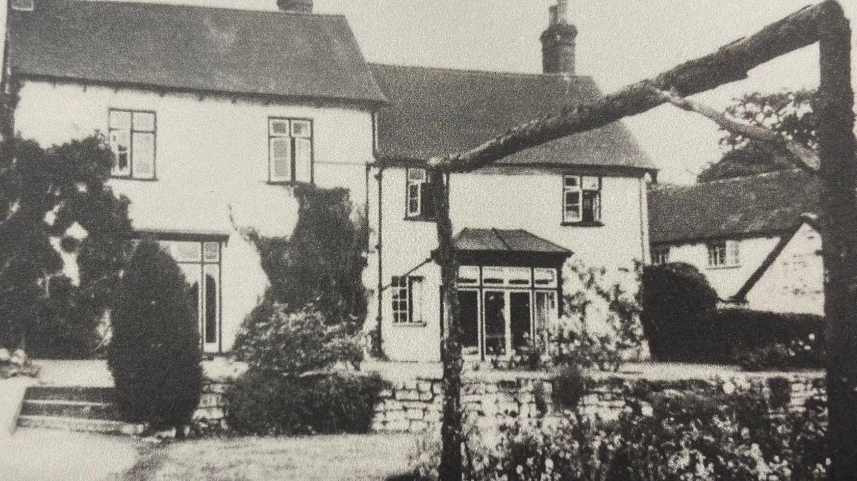 A black and white image of an old white wall building from its back garden with black framed windows and dark sloping roofs. A patio area steps down into a garden, with flowers, shrubs and hedges.