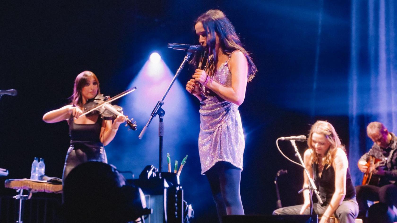 Éadaoin Ní Mhaicín is on stage at the Hydro. She is on the left of shot, playing her violin on her left shoulder. One of women from The Corrs is in the foreground playing a tin whistle. Another woman from the band and the male Corr are sitting to the right of shot listening to the music. The male musician is playing an acoustic guitar.