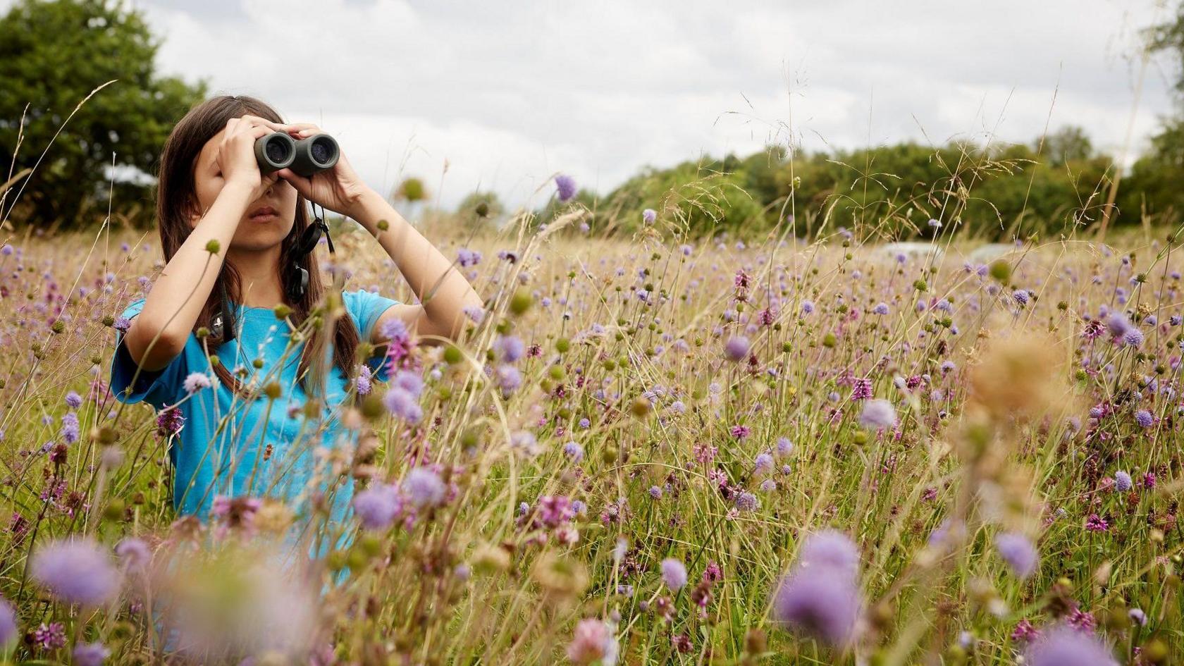 Mya-Rose stands in a field with purple flowers. She is holding a pair of binoculars up to her eyes as she spots birds in the distance. She is wearing a blue top and has long brown hair.