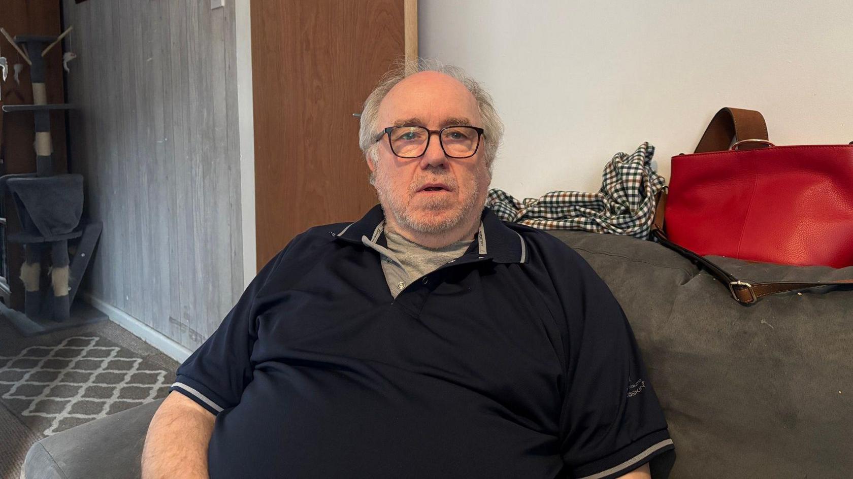 A man is sitting on a grey sofa. He has grey hair and back glasses and grey stubble. He is wearing a black t-shirt