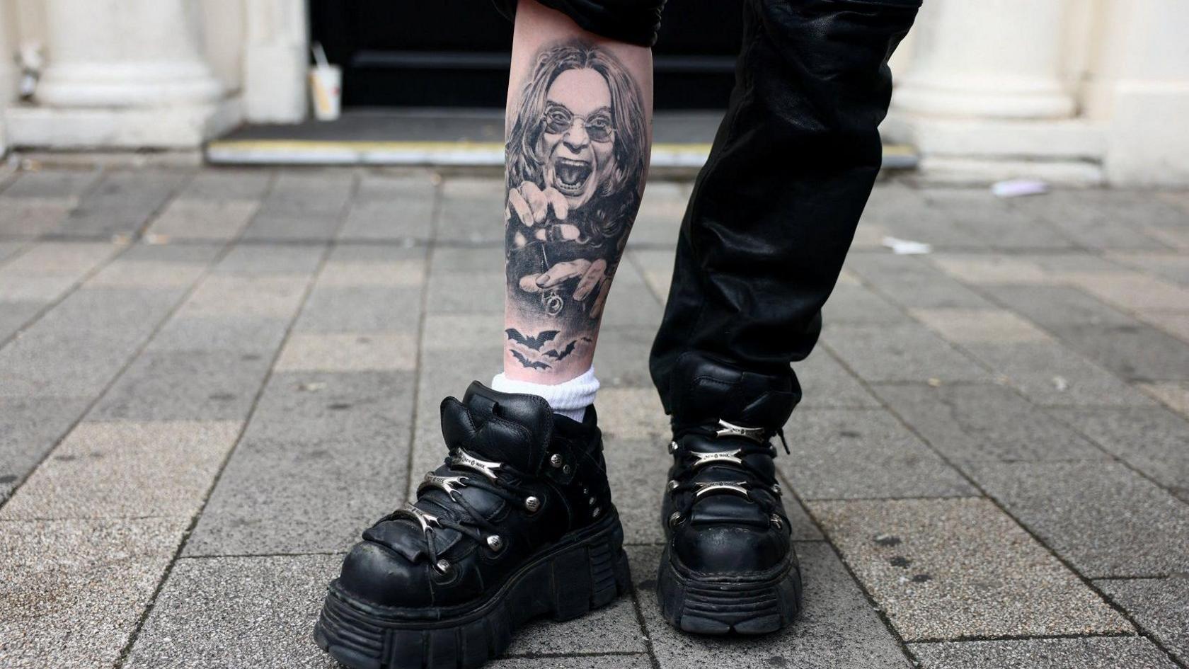 A person is wearing black platform boots and black trousers. One trouser leg is pulled up, exposing their leg with a tattoo of Ozzy Osbourne, who has long black hair and circular black glasses. There are bats underneath him and he has his hands out with his teeth bared