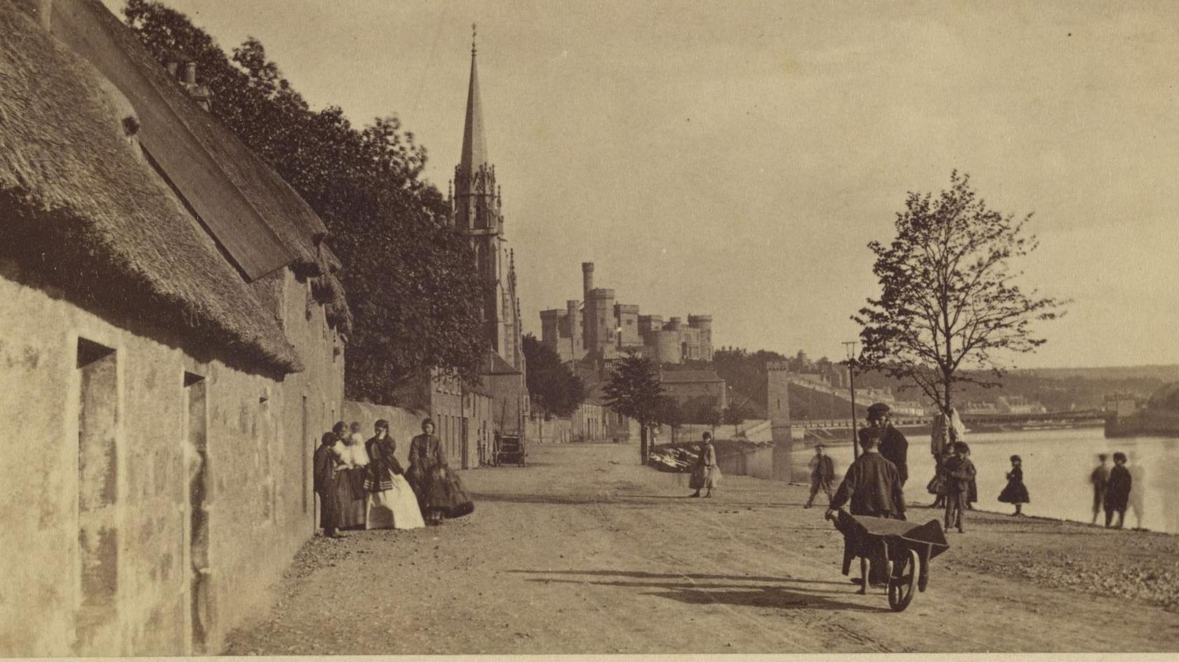 The sepia image shows an Inverness Riverside scene. On the left are cottages with thatched roofs. A group of women in long dresses are gathered near one of the cottages. They are standing on a rough, dusty road. A number of other people are on the road, including a boy who is pulling a wooden wheelbarrow and is talking to a man with a bushy beard and is wearing a flat cap. The River Ness flows on the right of the image and in the distance is a bridge and Inverness Castle.