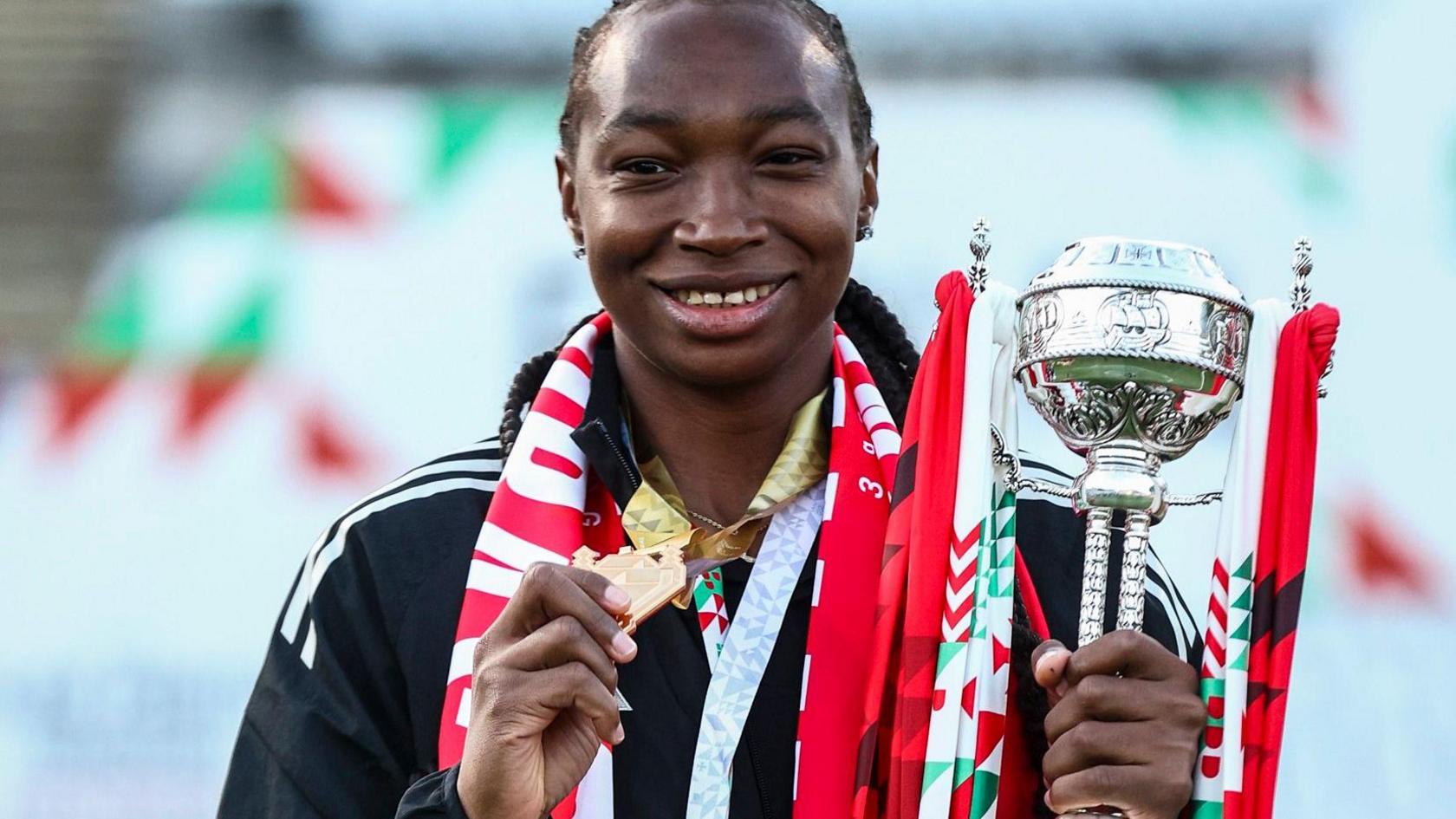 Michaely Bihina, visible from chest up, smiles as she holds up a golden medal which is hanging around her neck with her right hand as she holds a silver trophy with red and white ribbons attached to it with her left hand. She wears a black tracksuit top with a red and white scarf around her neck
