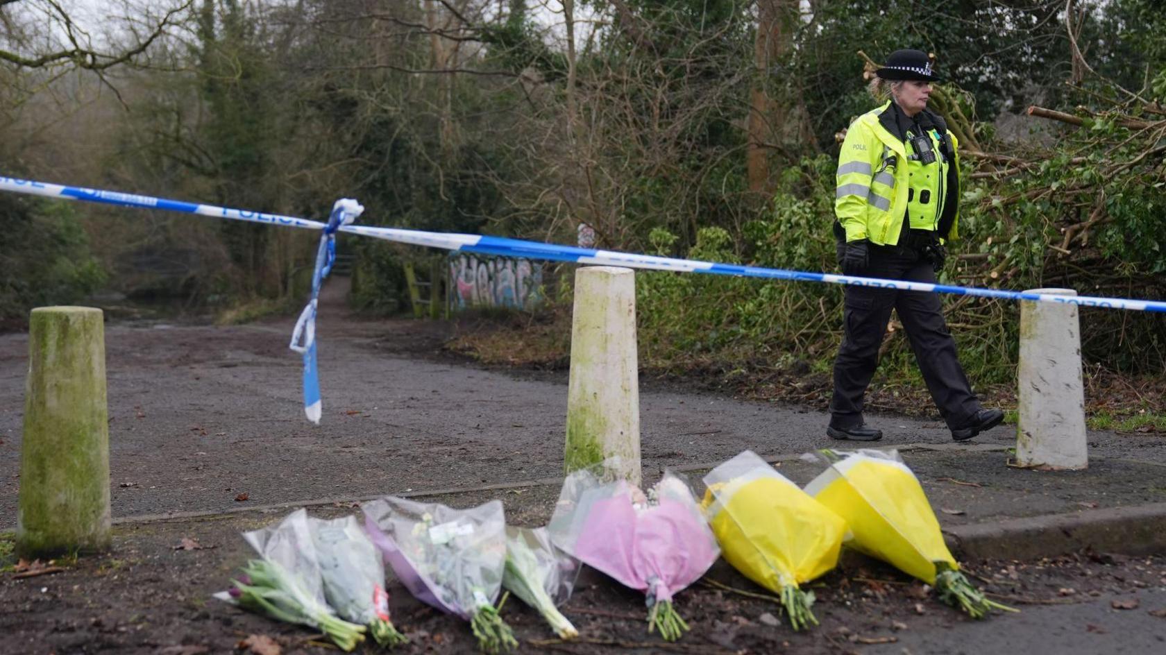 Bouquets of flowers are laid in a row on the ground in front of a police cordon. The flowers are wrapped in clear, pink and yellow plastic. Behind the cordon there is a muddy path lined with trees. A policewoman in a high-vis jacket is walking behind the cordon.