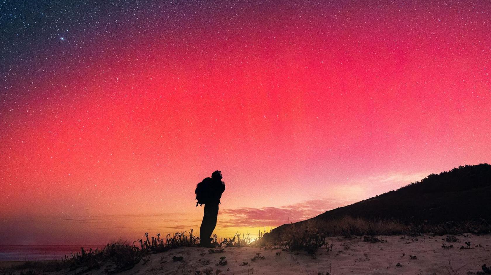A person wearing a backpack looks up at red northern lights in the sky.