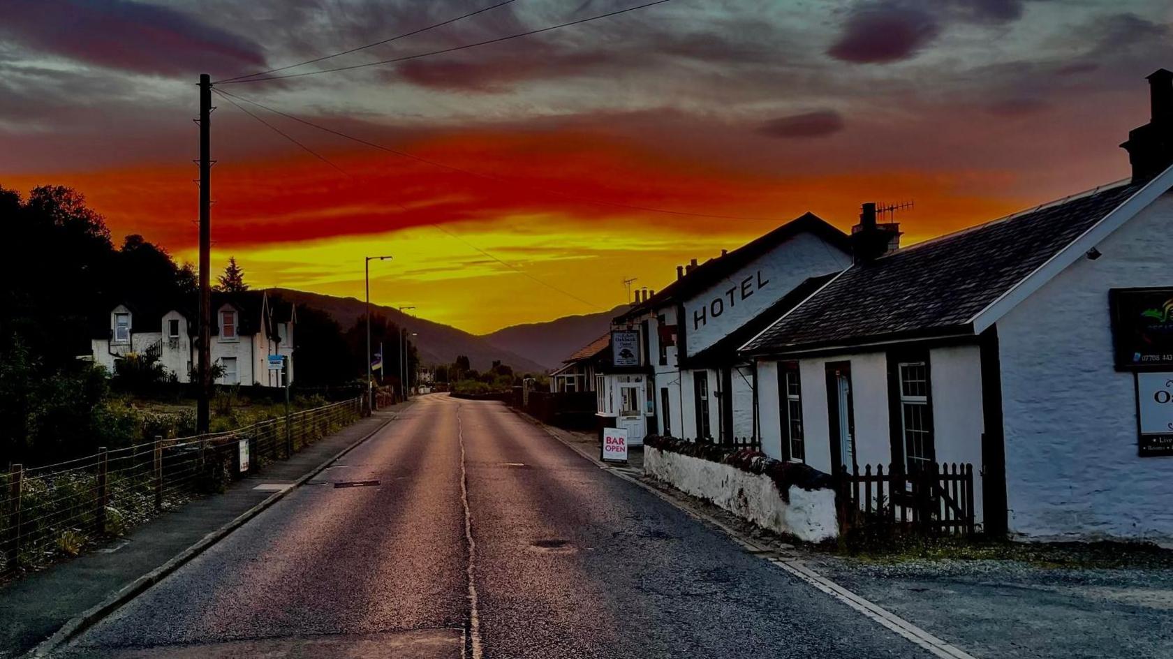 An orange and yellow sunset illuminates the street of the Inn. The road runs down the middle of the picture as the hotel sits to the right. Its a white, quaint building. It looks like a cottage and has the letters hotel printed on the side. 