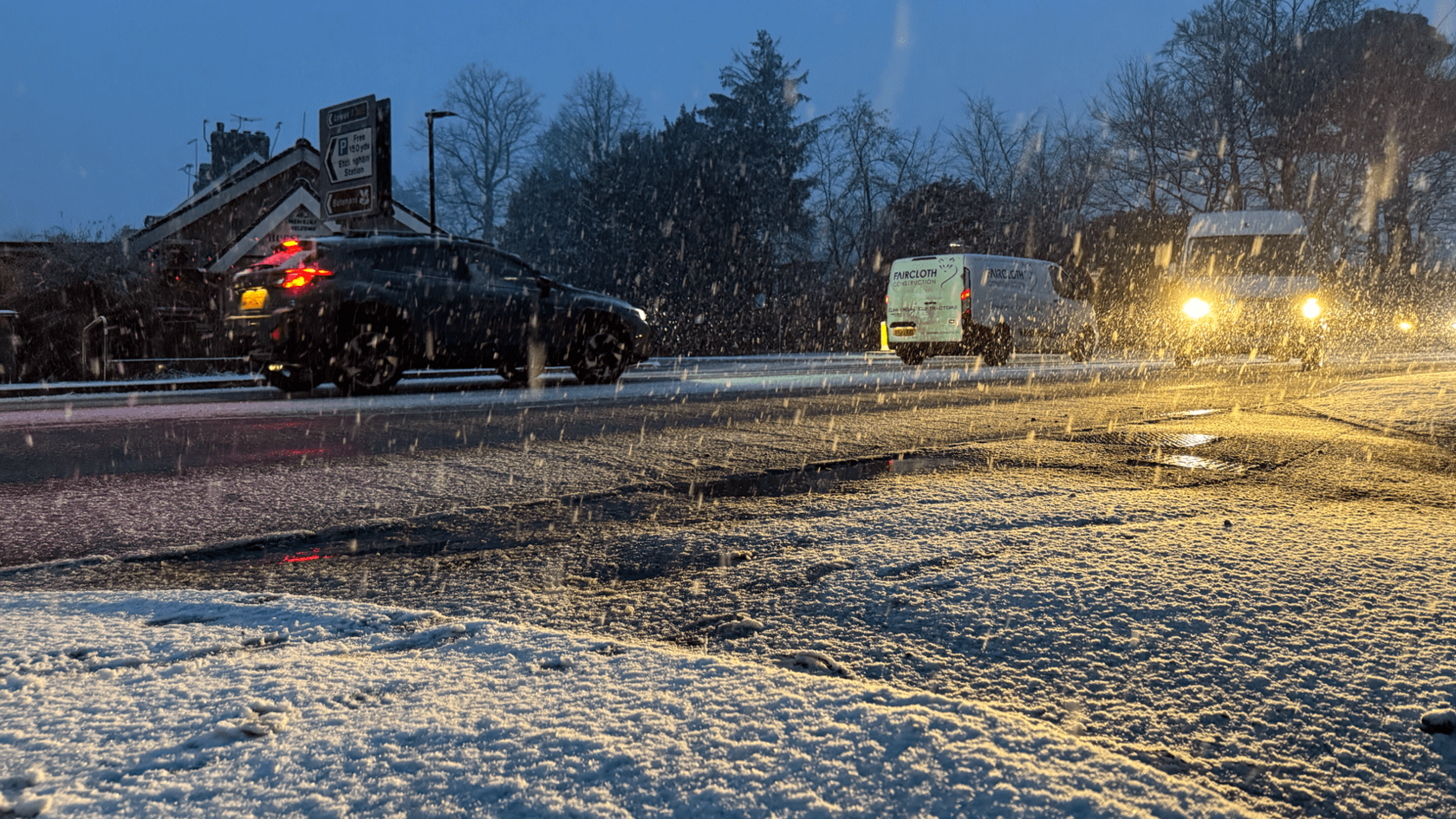 Snow falls on a main road as two vans and a vehicle drive past