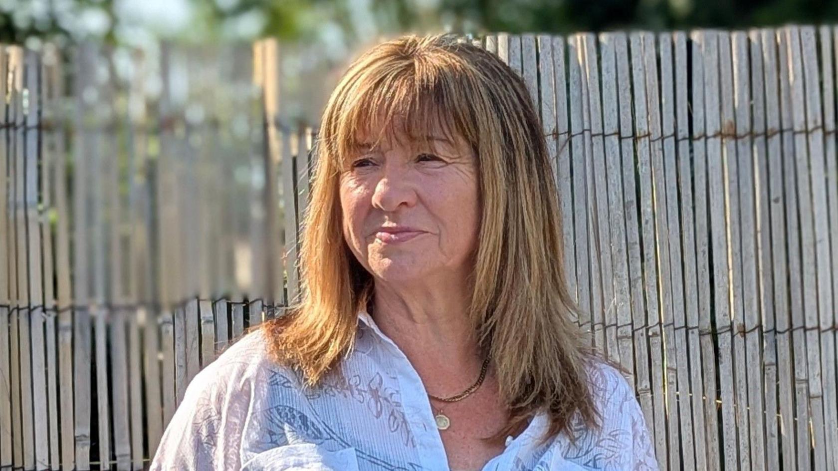 A head and shoulders photograph of Kathleen Richards, wearing a white blouse with flower detail, looking off to her right. She is standing in front of a bamboo style high fence and it is a bright day.
