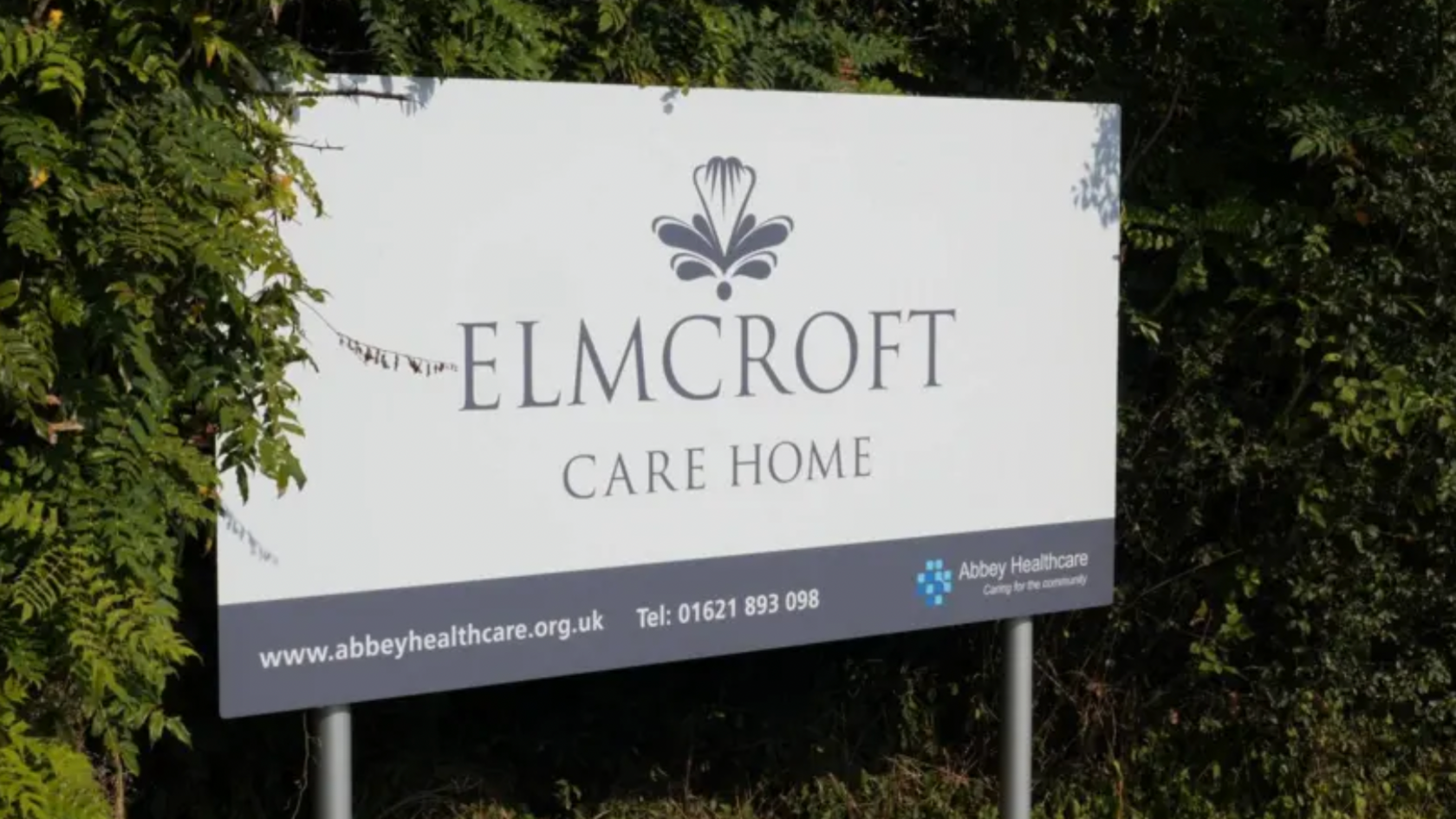 A sign that reads Elmcroft Care Home, installed in front of bushes or trees.