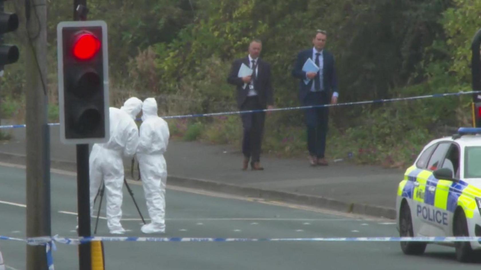 Forensic investigators in white overalls behind blue and white police cordon, with other detectives in background and police car to one side