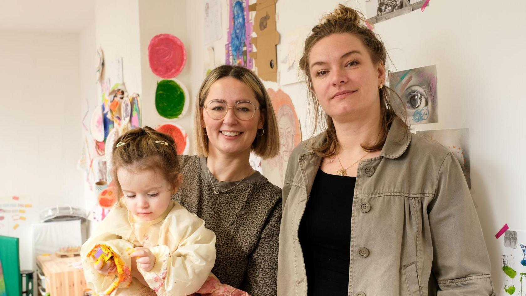 Two women stand close together in a room that has artworks on the walls behind them. The woman on the left has chin-length blonde hair and glasses, and she is holding a toddler who is wearing an art overall. The woman on the right has brown hair that is tied up and is wearing a black top and a grey shirt.