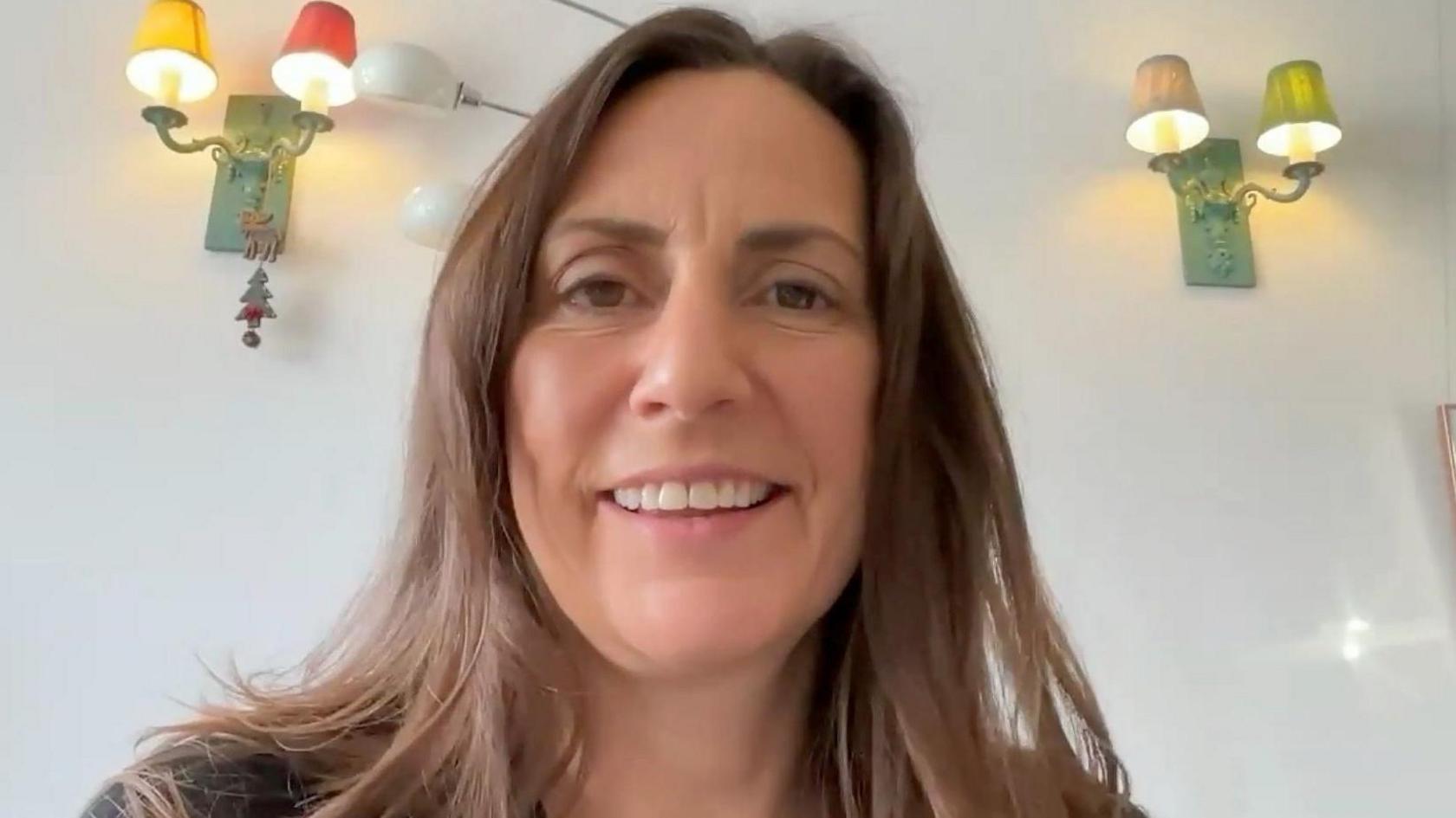 A woman with long brown hair, talks to camera from home, there are wall lights in the background. 