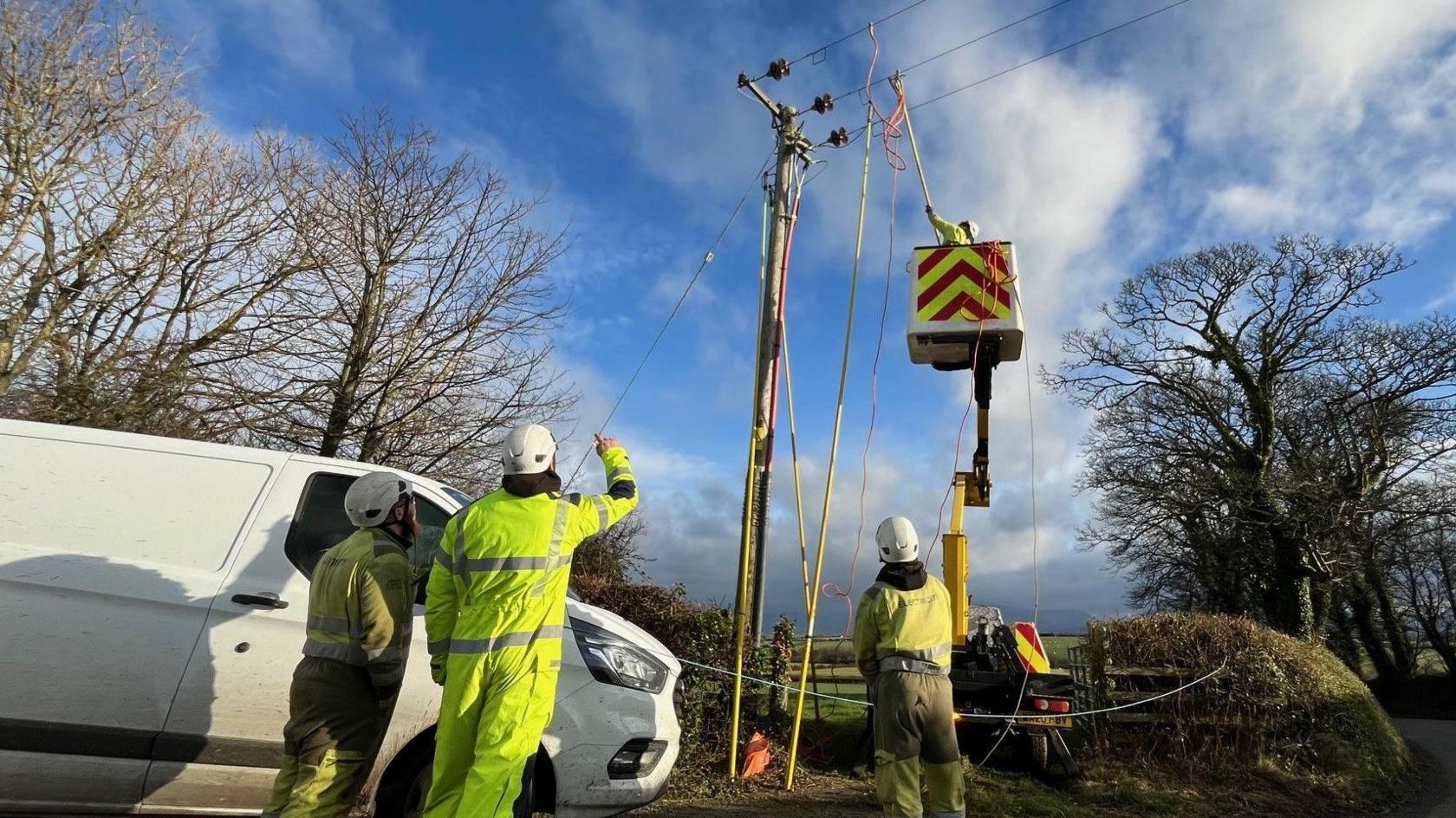 three workers in high viz clothing standing beside a white van parked beside a telegraph poll which has a cherry picker beside it and another worker reaching up towards a the wires