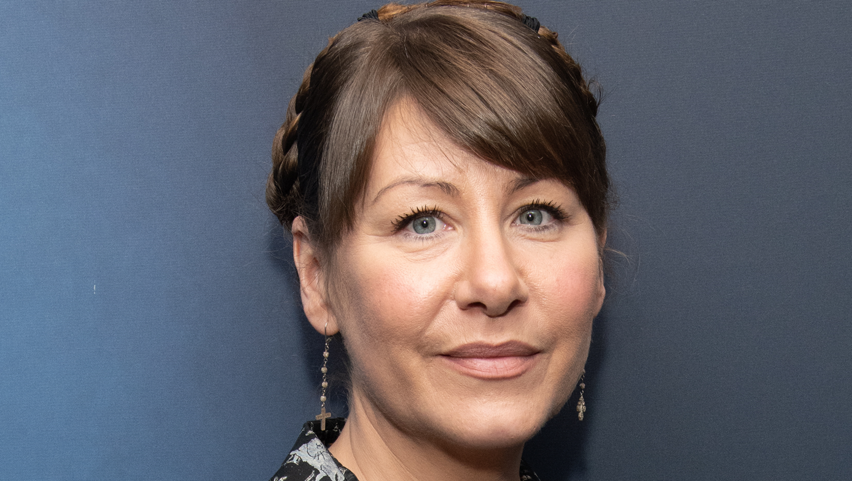 Rachel Fuller, who has brown hair tied up and is wearing long earrings, standing in front of a blue wall. 