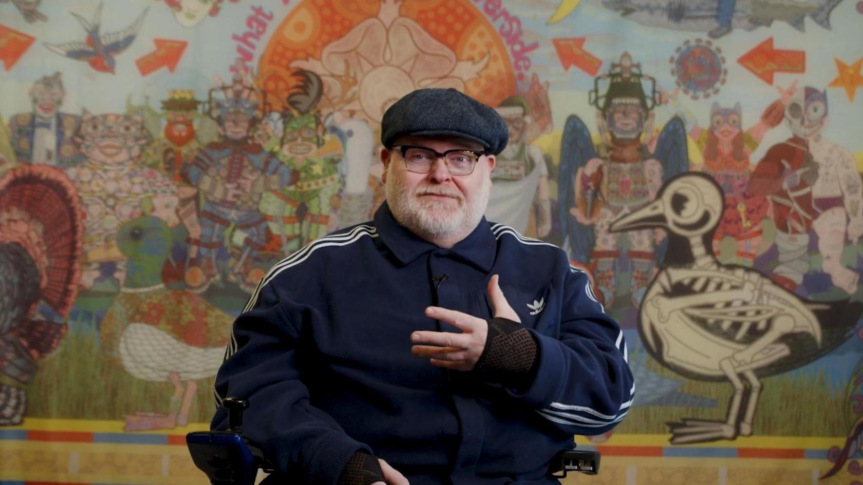 A man wearing a flat cap, a striped jogger style jacket and black glasses sits in front of a colourful wall or mural. He looks to be sitting in a wheelchair.