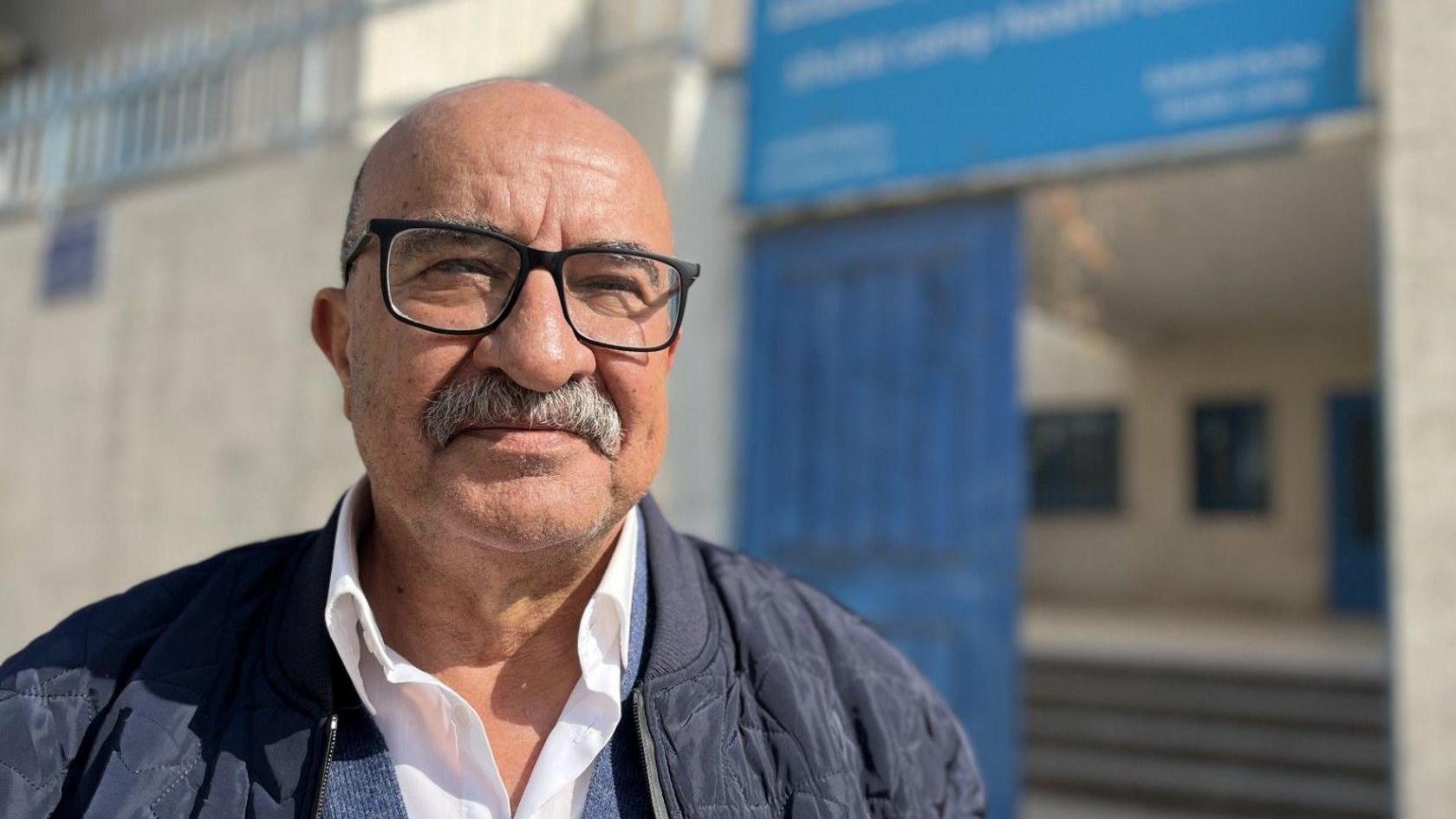 A bald man in glasses and with a moustache smiles at the camera, standing in front of a building with blue signs on the door