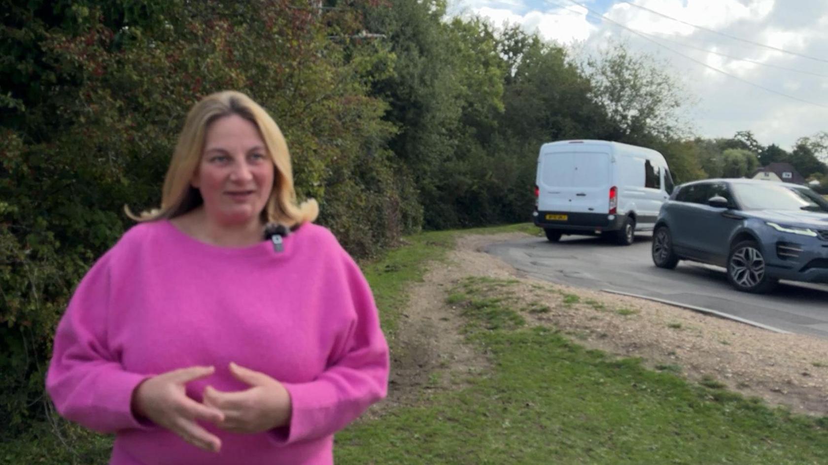 Hannah is wearing a pink woollen top. She is standing next to a road with cars going past behind her