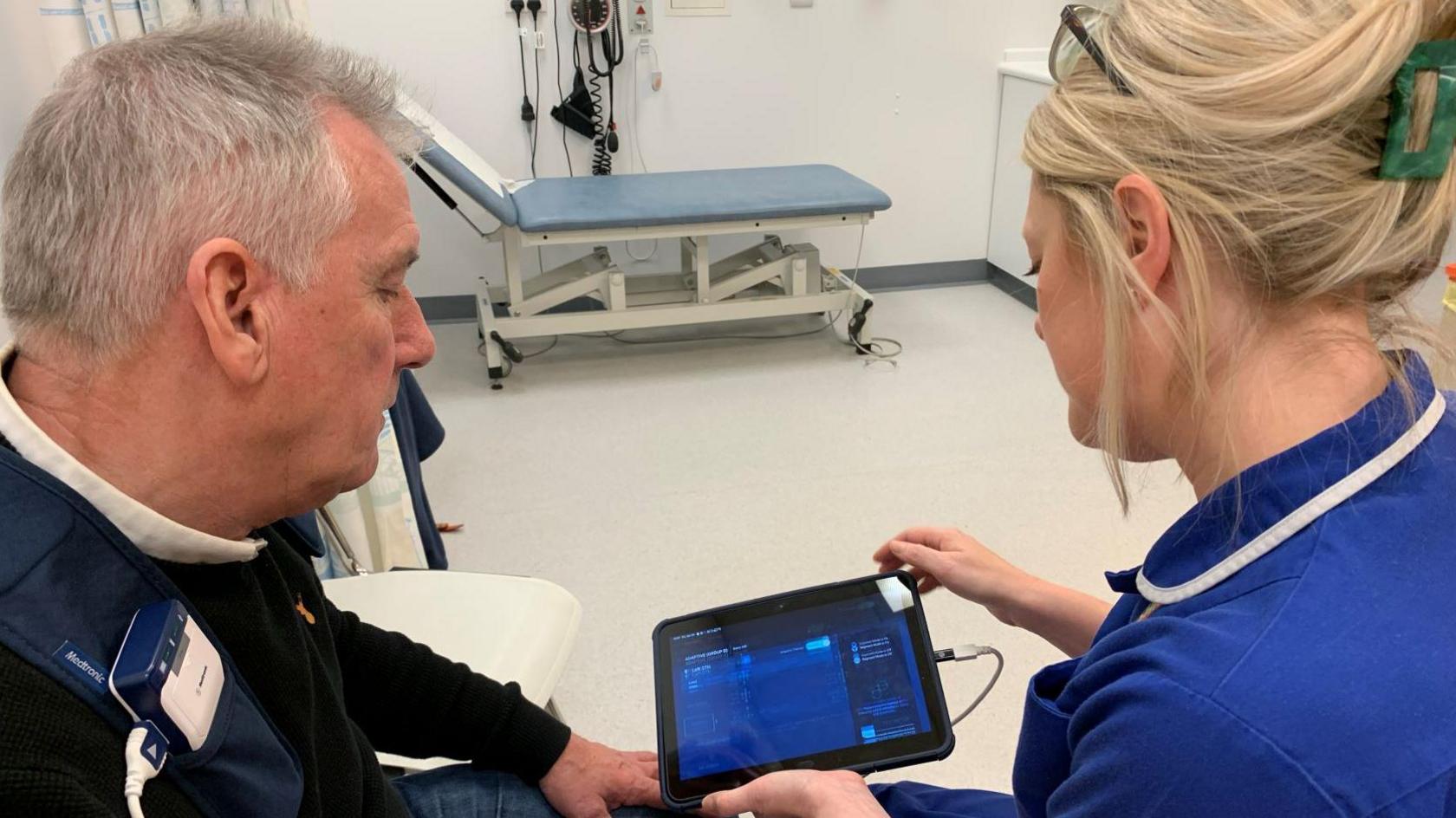 Kevin is with a nurse at hospital as the new system is re-programmed and switched on. They are both looking at a computer screen which is wired to his chest. 