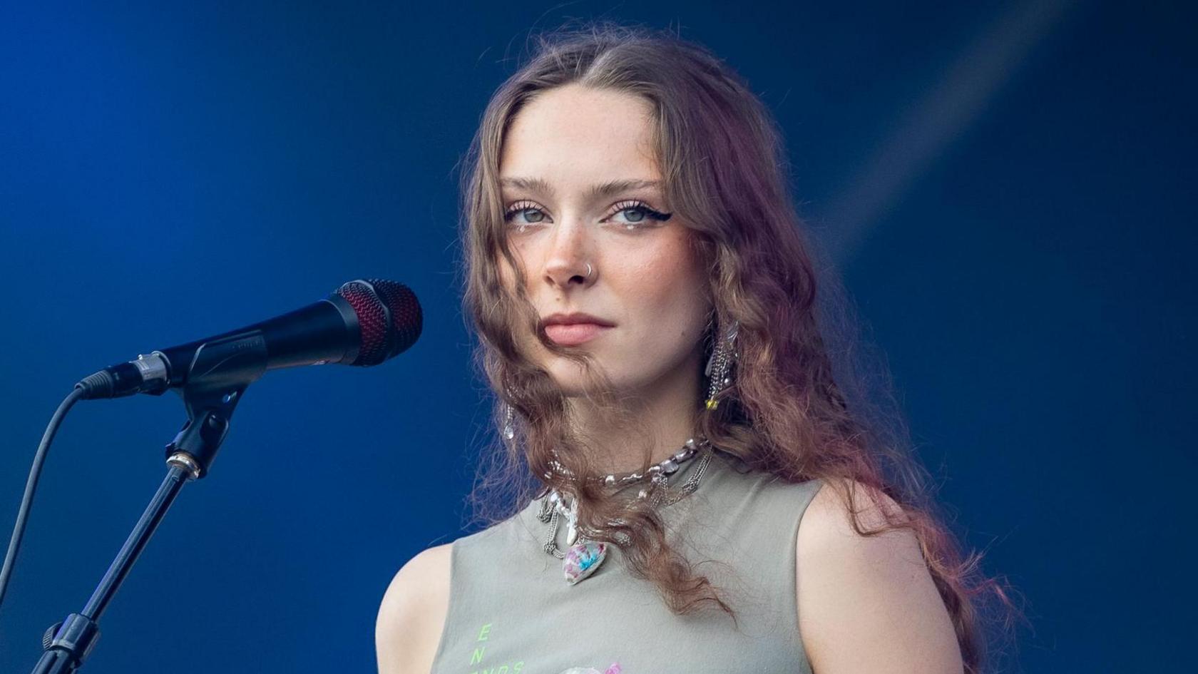 Holly Humberstone on stage. Holly's a 24-year-old woman with long curly brown hair worn loose. She has blue eyes, rimmed with a thick black eyeliner. She wears a high-necked sleeveless grey top with a chunky silver necklace and is pictured against a blue backdrop.