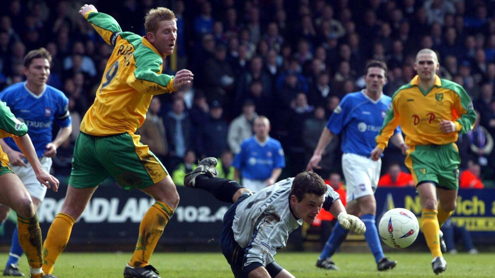 Andy Marshall is wearing a grey Ipswich top as he dives towards the ball at Carrow Road. He is surrounded by players either wearing the blue outfield strip of Ipswich or the green and yellow of Norwich. A busy crowd watches on from the stands.