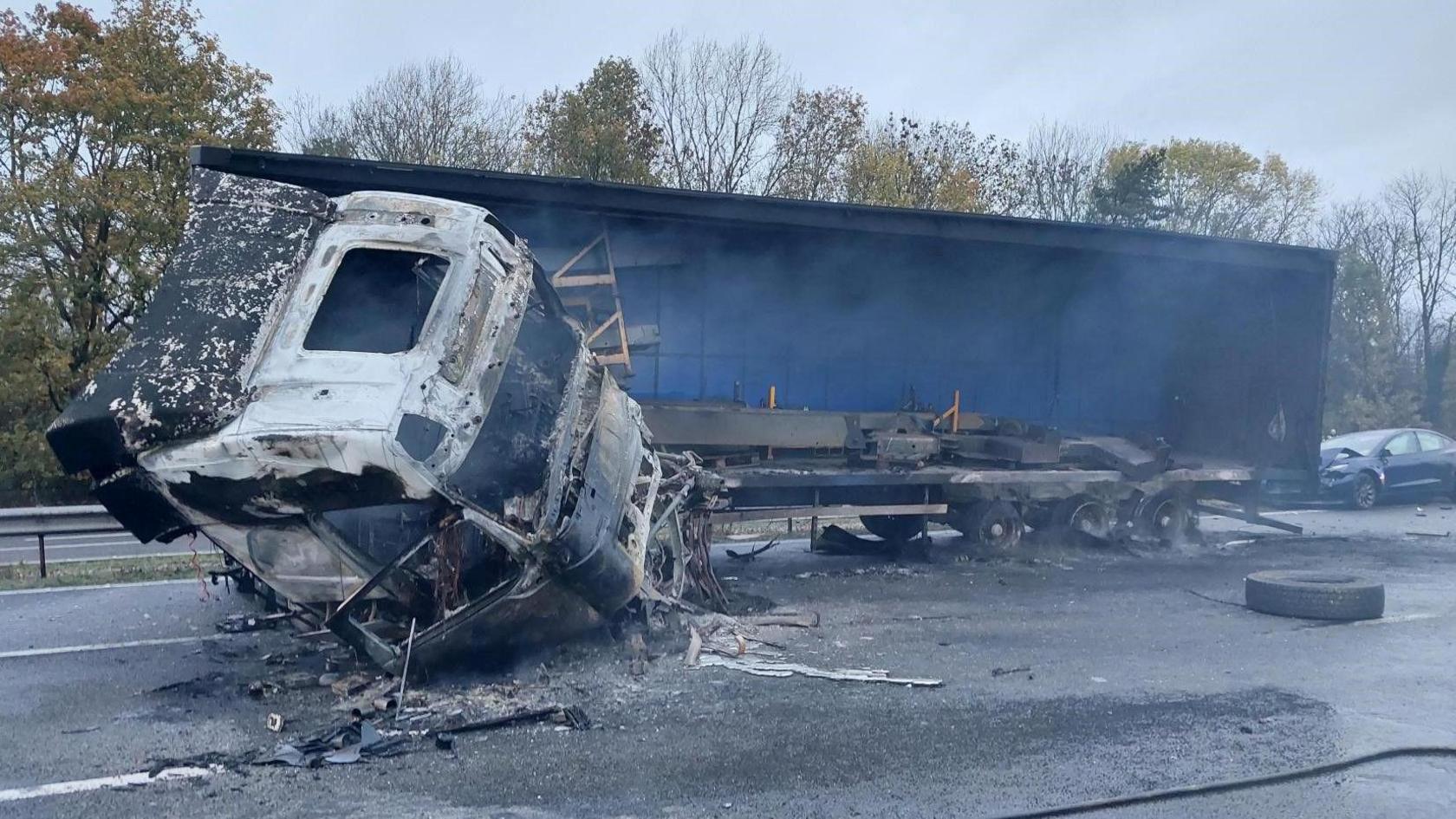 A burnt out lorry and trailer that's jack knifed on the motorway. The road is severely damaged and debris surrounds the vehicle