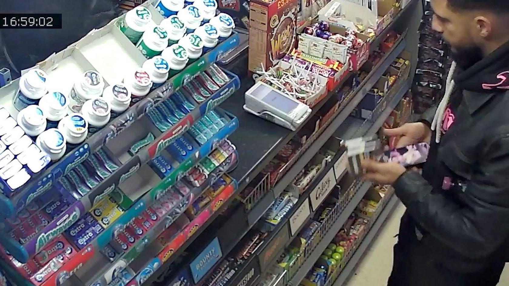 A still image taken from CCTV footage. It shows Mohamaddi at the till of a convenience store clutching some cigarettes and a phone he had used to purchase them. His face is barely visible and he is wearing a black hoodie with pink writing, covered by a black jacket.