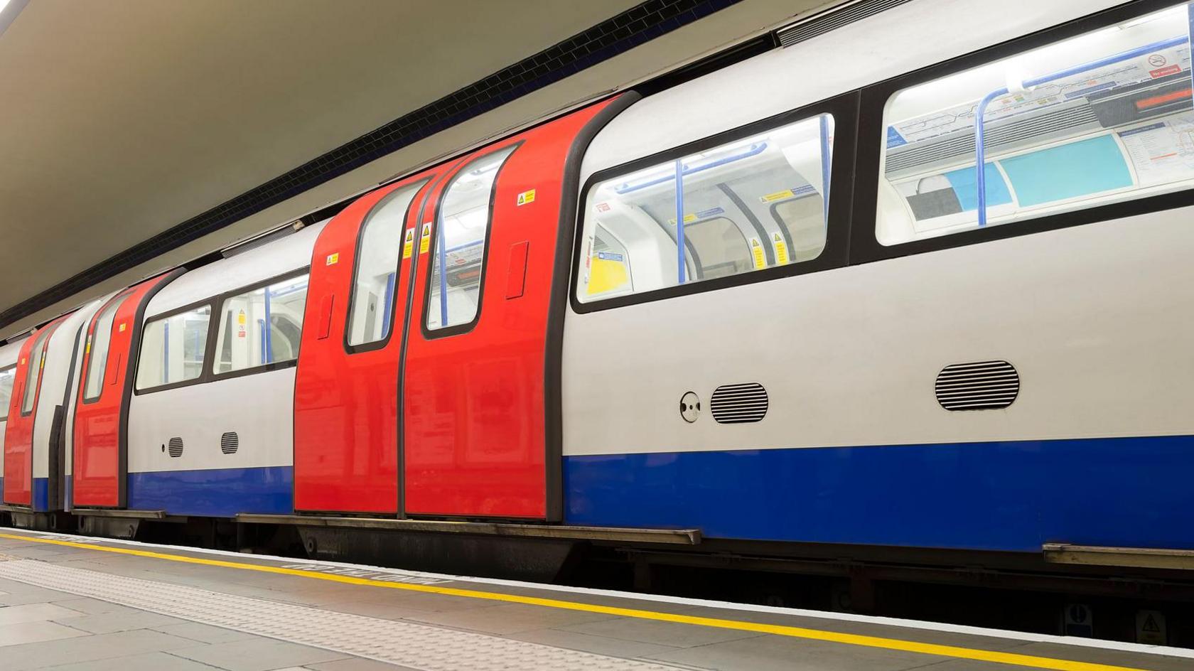 A London Tube train in white and blue livery with bright red doors, at the platform in an underground station. 