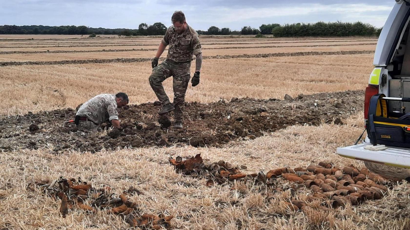 Two men in camouflage Army uniform are looking at bombs in a pit in a field.
There are a number of the bombs lined up behind a police vehicle, only part of which can be seen. The field is brown and stubbly