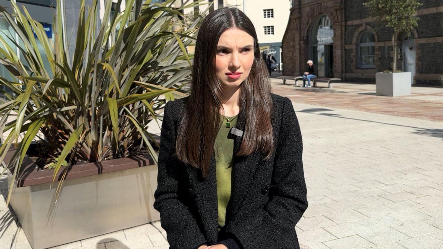 Lisa Creevy sits in a sunny town square. She has long brown hair and is wearing an olive green top with a block blazer-stye jacket over it.