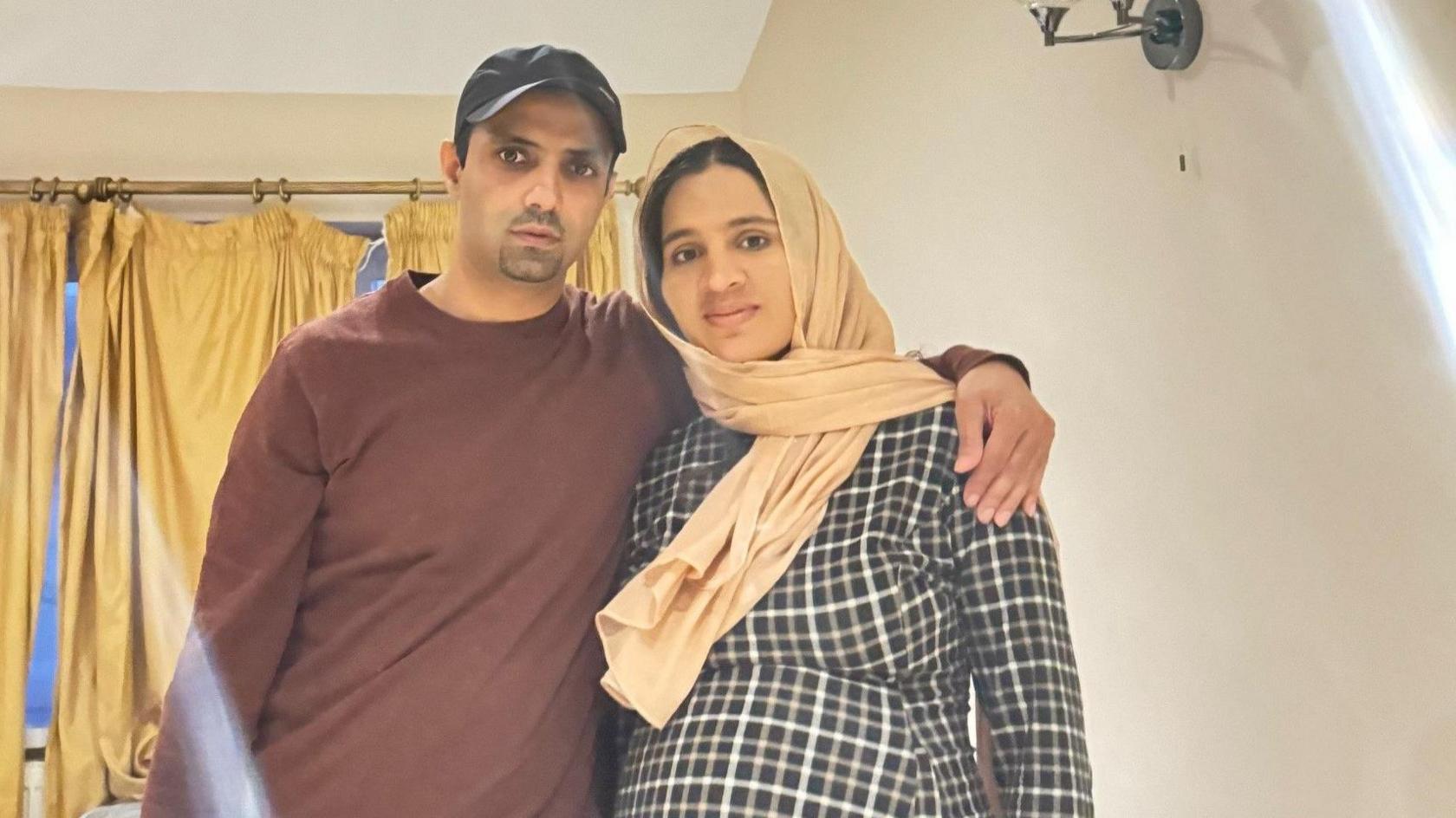 Nabeel (left)  and his wife (right) Nabeel is wearing a brown long sleeve t-shirt and his wife is wearing a checked dress.