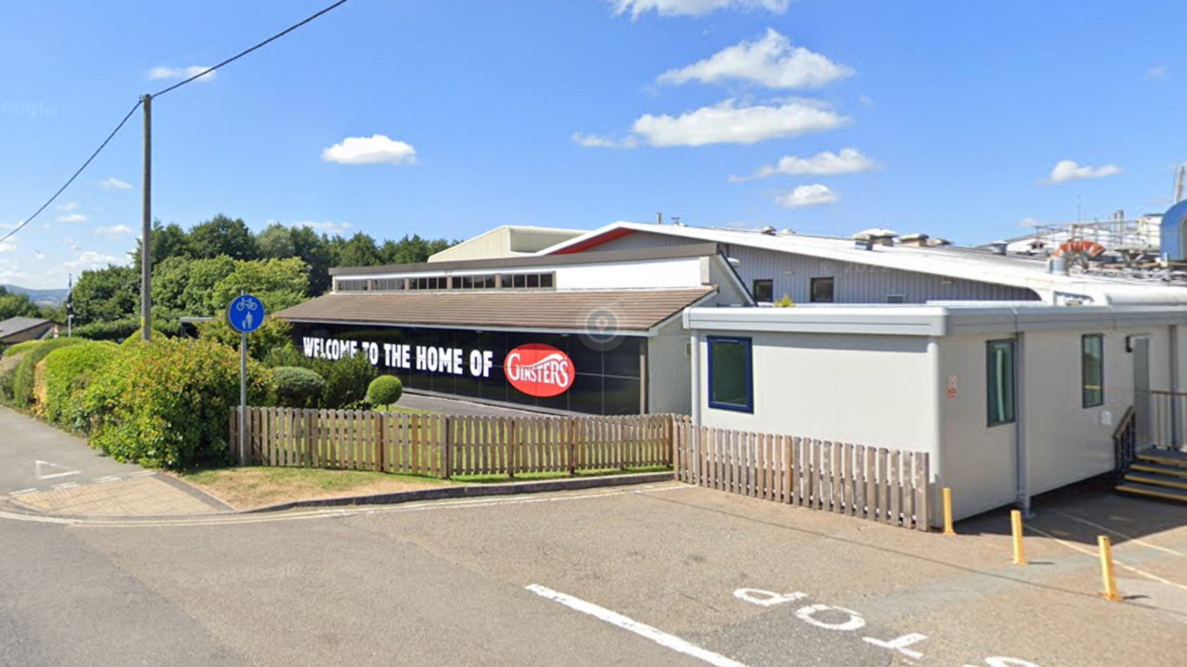 A pale coloured single storey industrial unit by a road with the words welcome to the home of Ginsters.