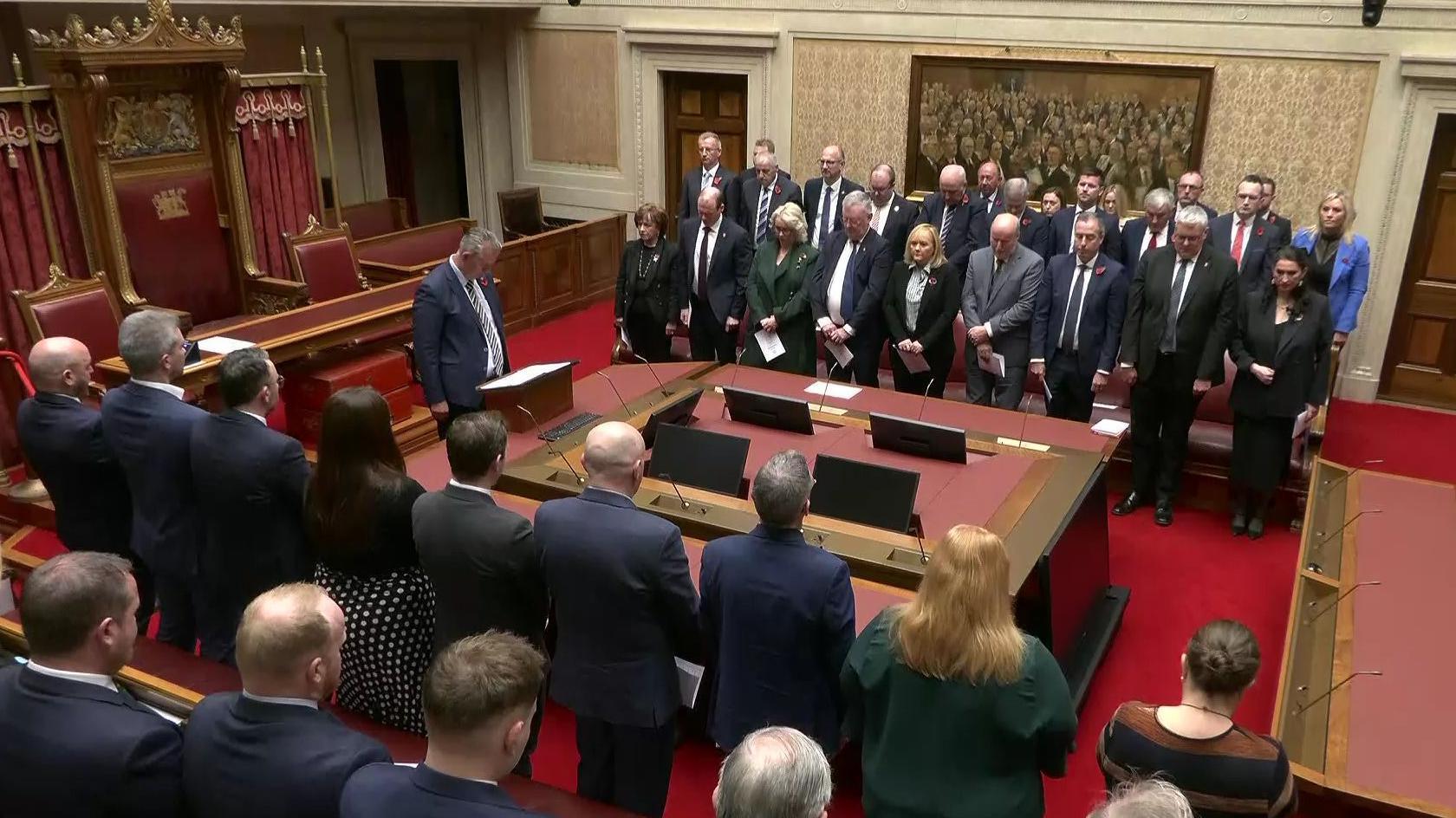 Assembly members stant in silence in Stomont's Senate Chamber during an Armistice Day ceremony.  Speaker Edwin Poots, who led the ceremony, is standing at a lectern in the middle of the room with his head bowed