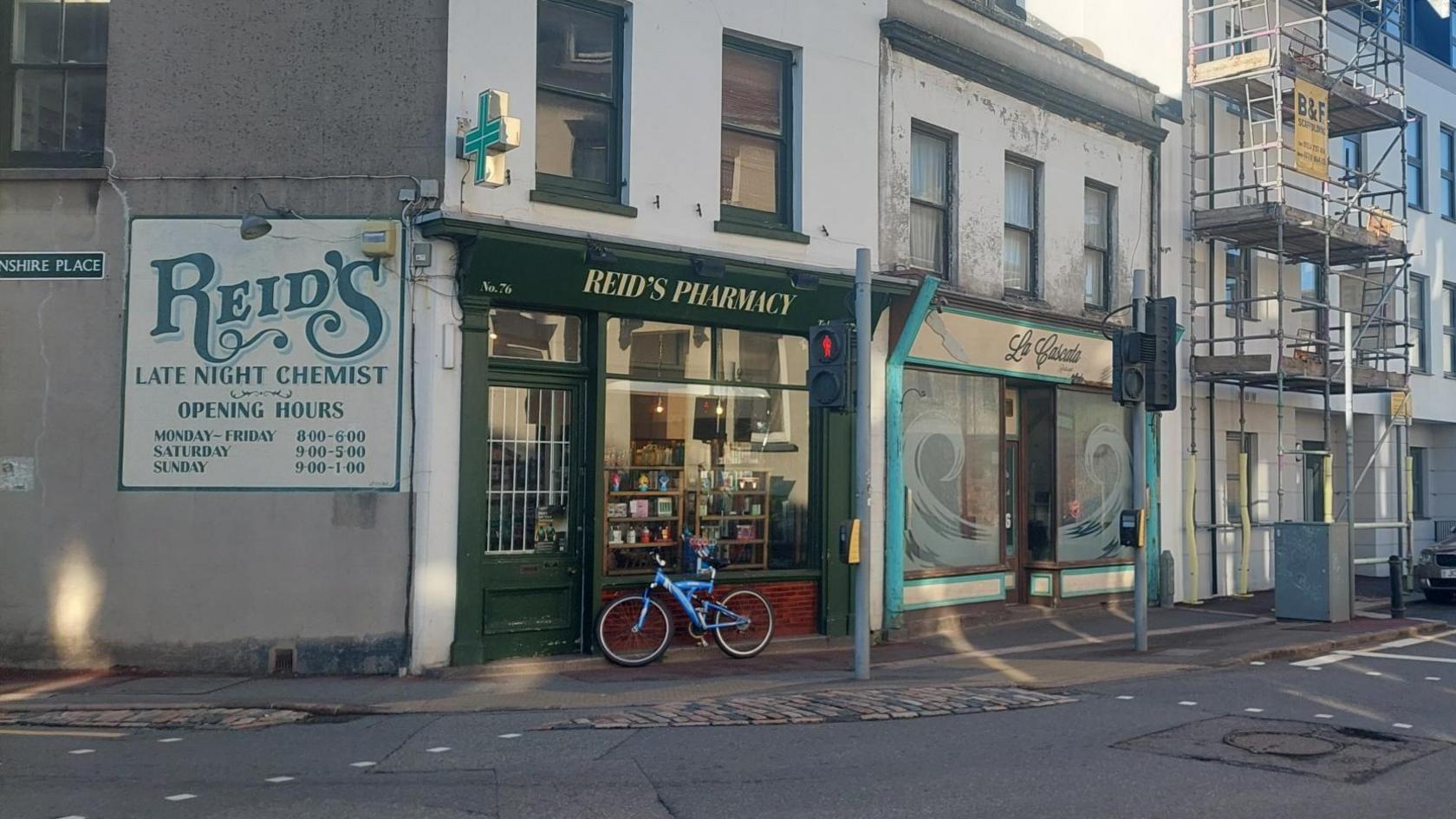 The outside of Reid's Pharmacy in Jersey. It is on the corner of the street and is painted green with cream writing. There is a bike left outside. There is also a pedestrian crossing.