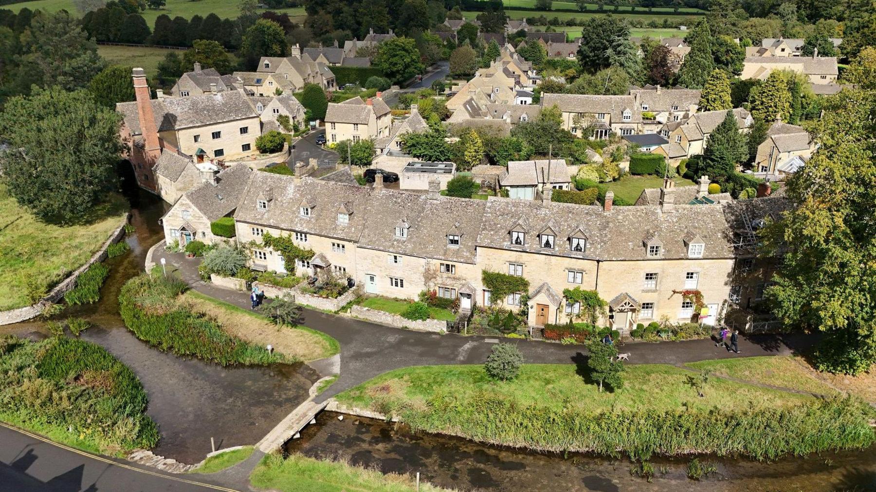 An aerial shot of Cotswold village Lower Slaughter, taken from a drone. The moat around the village edge is visible in the foreground, with the sandstone houses behind. The fields and trees ringing the village are still green