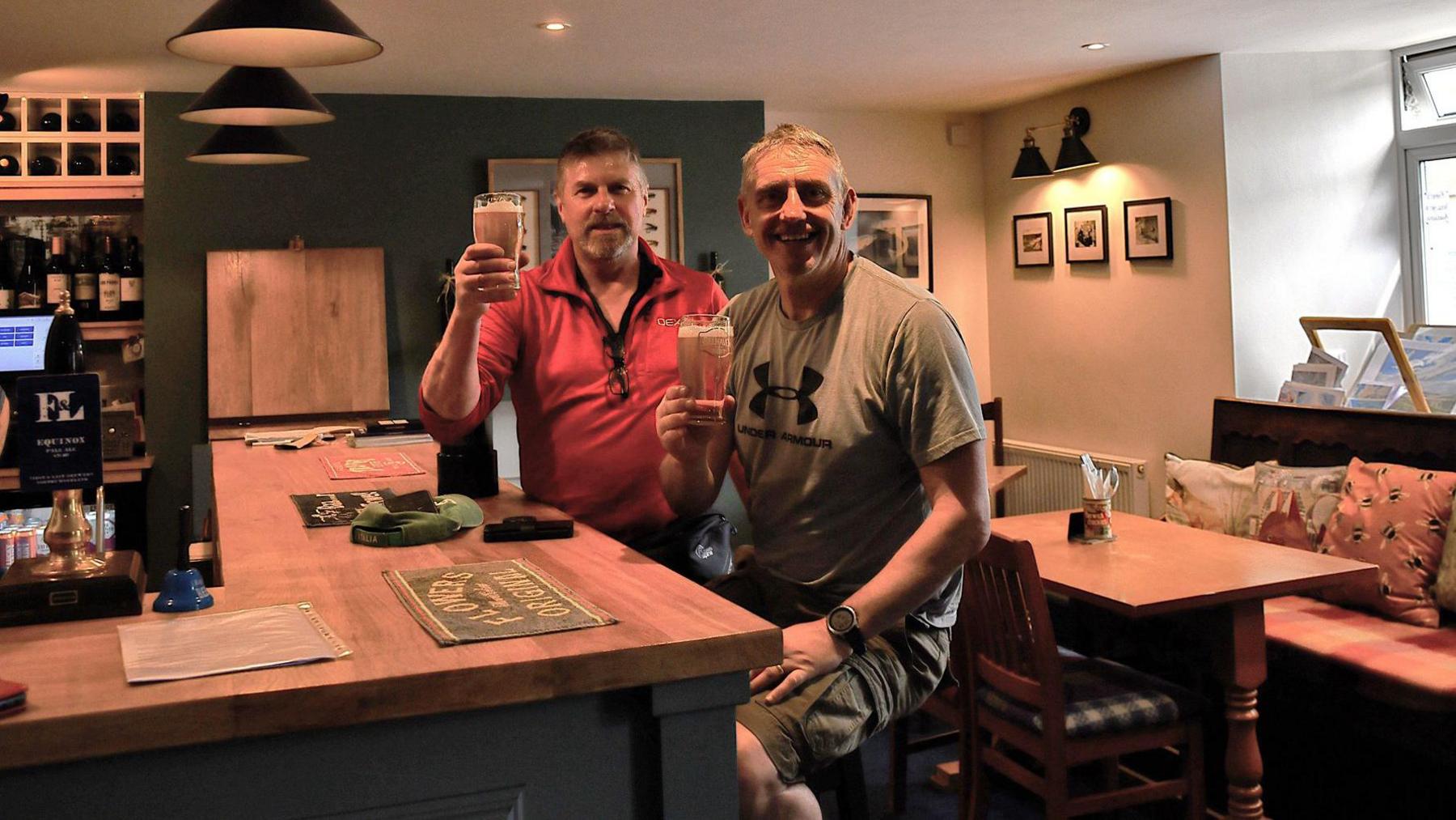 A man in a red t-shirt and a man in a grey t-shirt holding up a pint of beer inside a pub.