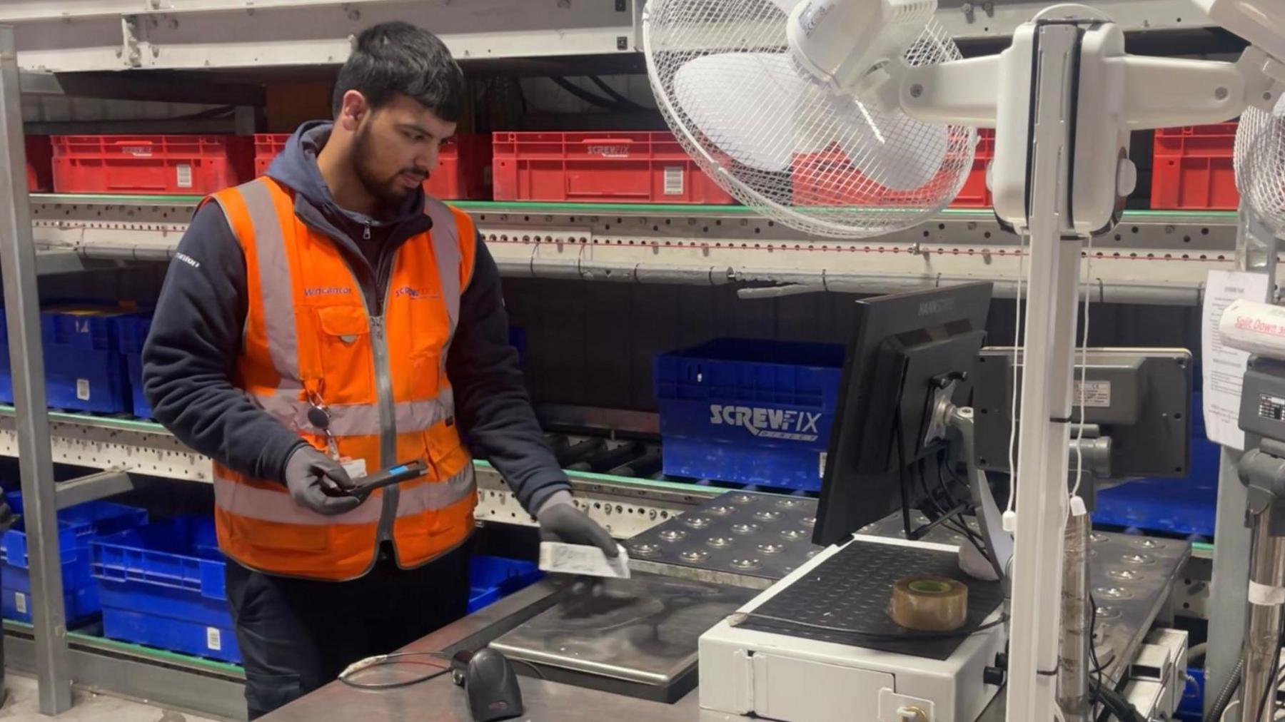 A teenage warehouse worker scans his pass at a computer beside a goods carousel.