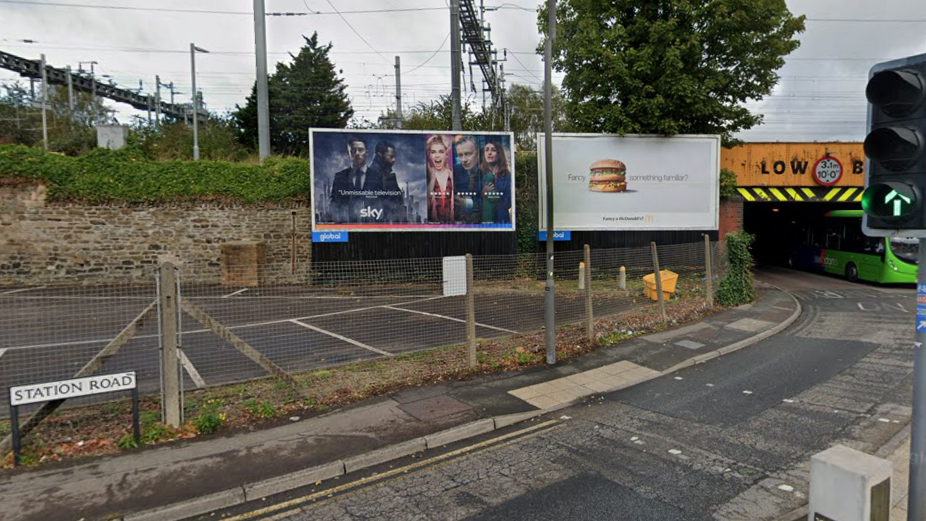 Station road in Swindon. The road bends around goes underneath a low railway bridge. There is a pavement, with a carpark to the left. 