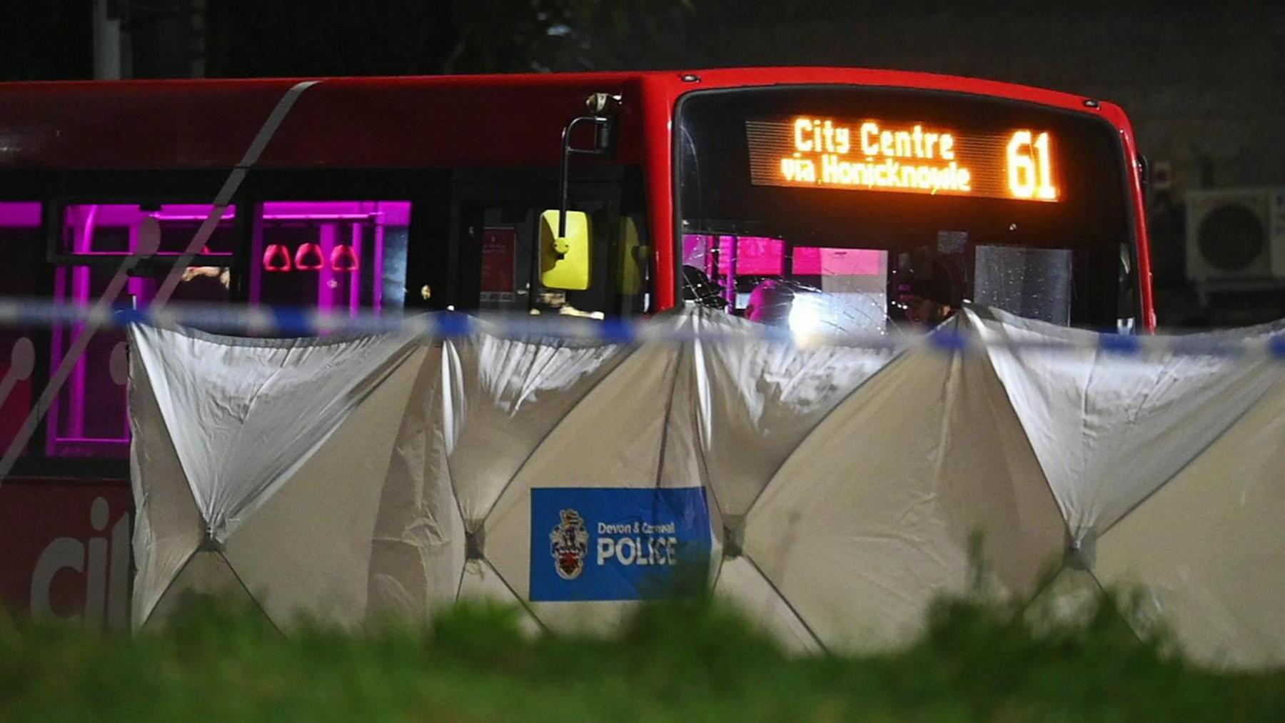 A red single-decker bus at night with a police investigation tent in front of it with the words Devon and Cornwall Police.