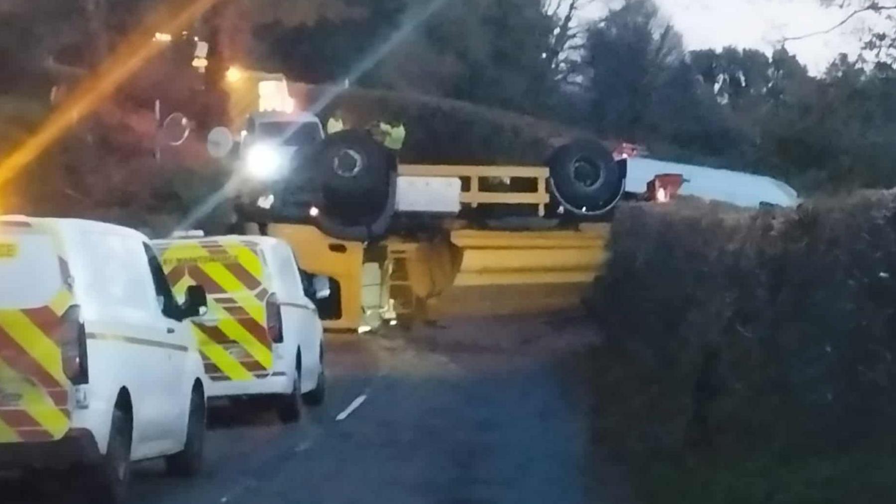 A yellow gritting lorry has overturned and blocked a road in Dartmoor. Two white vans are next to the lorry. Other vehicles with their lights on are on the opposite side of the gritter with people wandering around the scene.