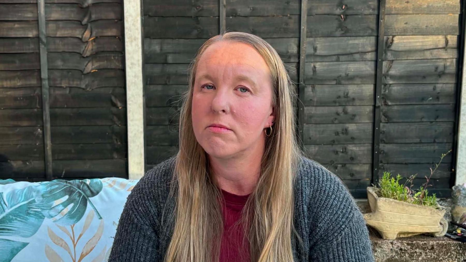 A woman sitting outside in front of a garden fence. She is looking at the camera with a serious expression. The woman is wearing a grey cardigan and a red t-shirt.