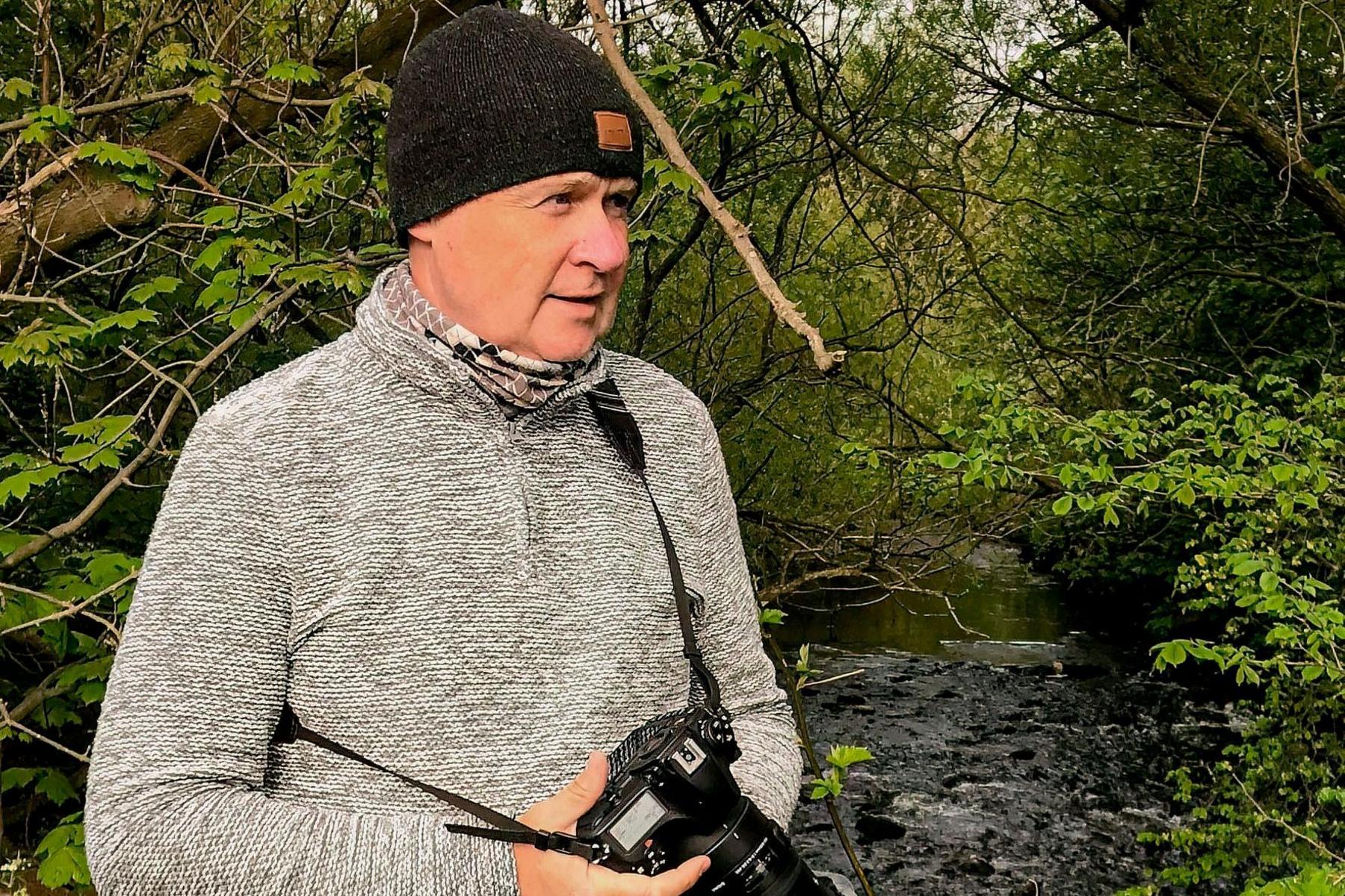 Tom is wearing a black woolly hat and grey jumper. He is holding a camera with a long zoom lens and is standing beside a river with tree branches over head.