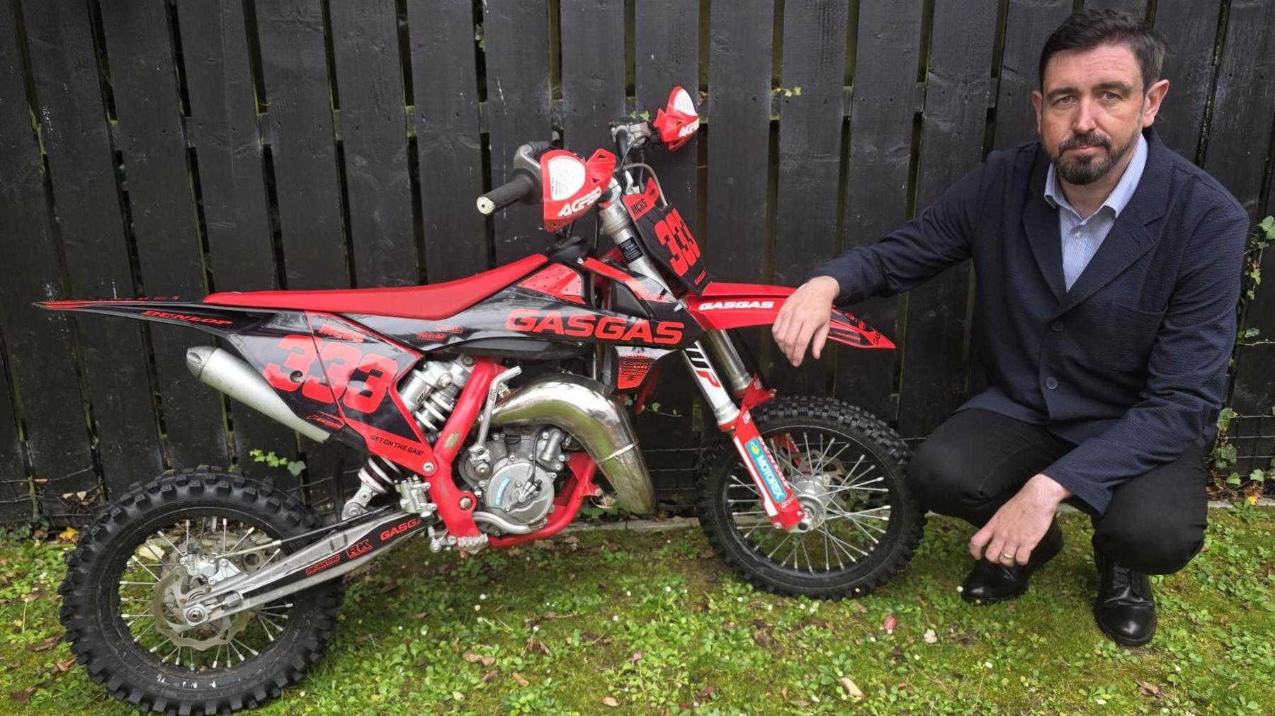 Paul Doherty crouched down beside a red scrambler type bike. He is wearing a navy blazer and blue shirt with black trousers and shoes.