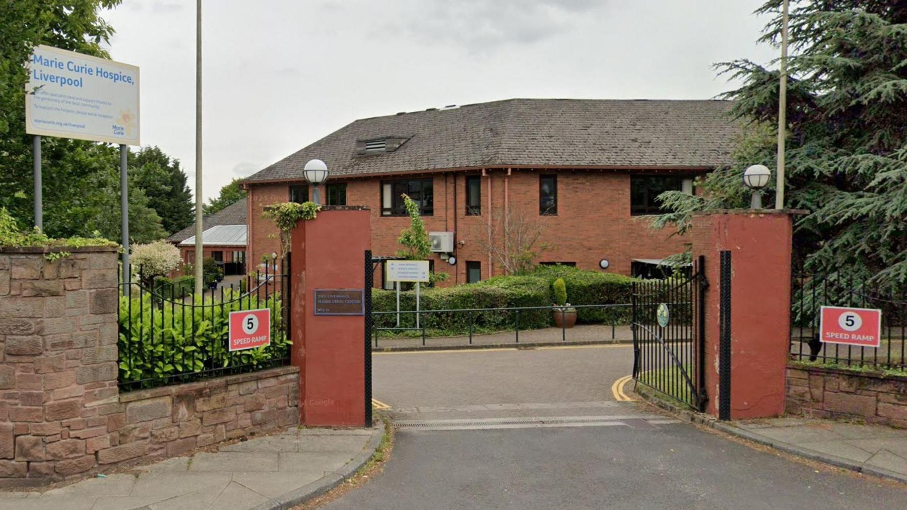 A red brick building set behind a sandstone entrance and green trees and bushes