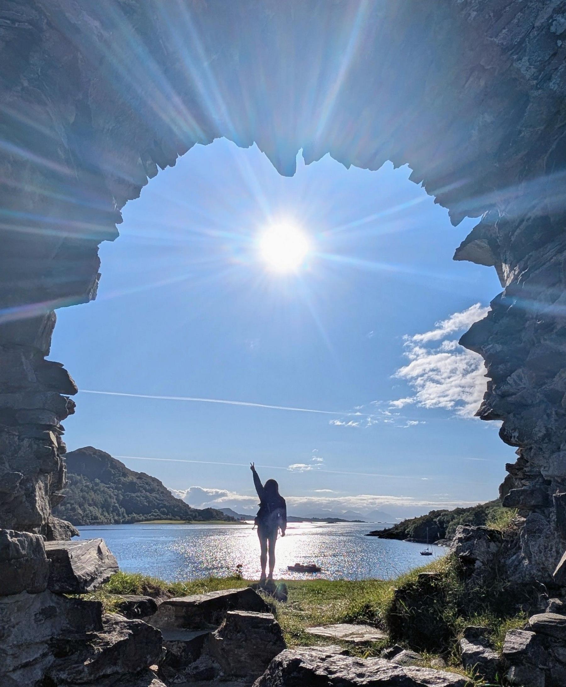 We look through a stone archway to see a woman standing on a rock, the loch behind her, the sun shining on the water and in the sky, dead centre under the arch.
