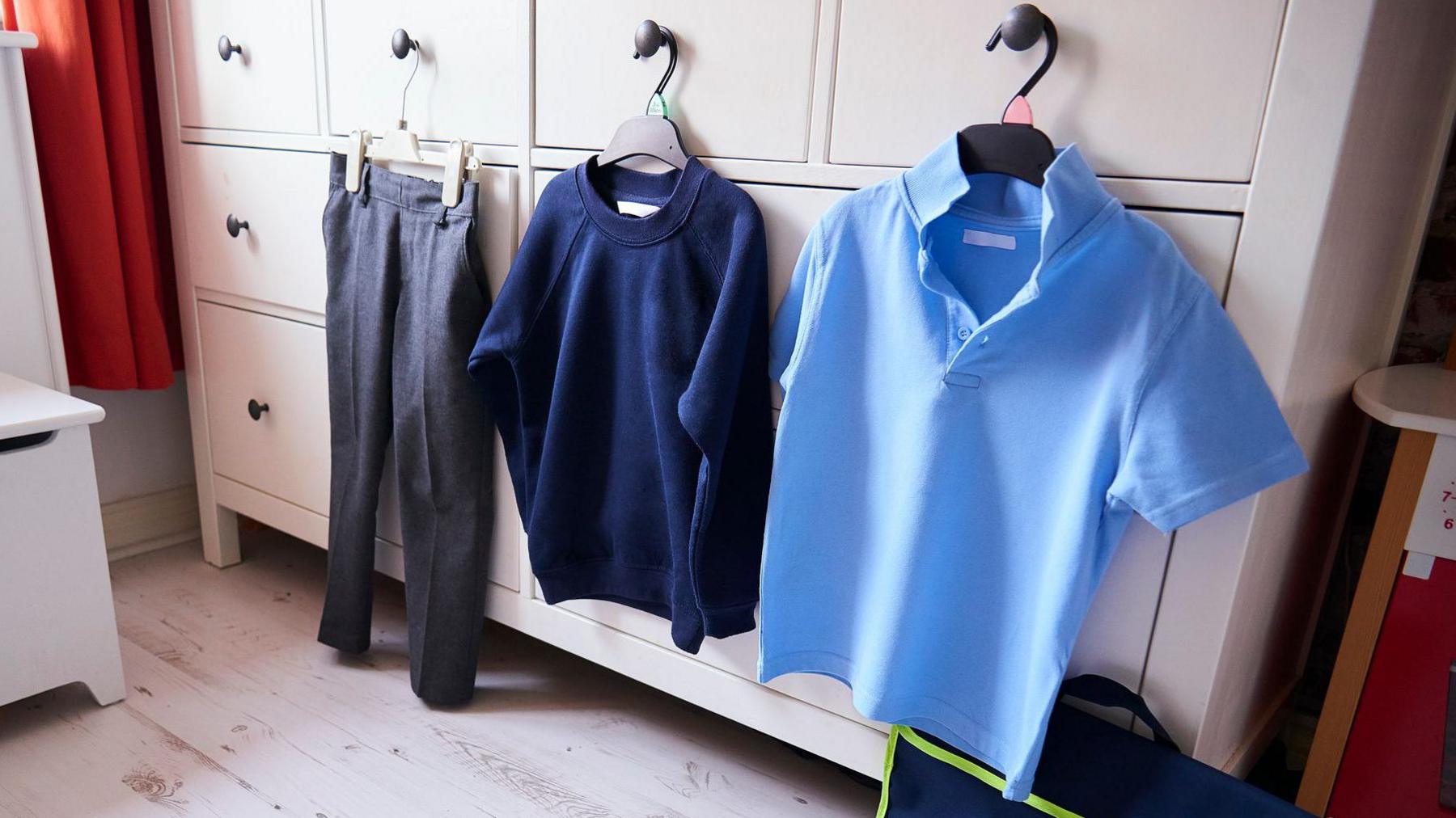 A white chest of drawers with a child's grey pair of trousers, navy jumper and blue polo t-shirt hanging on it.