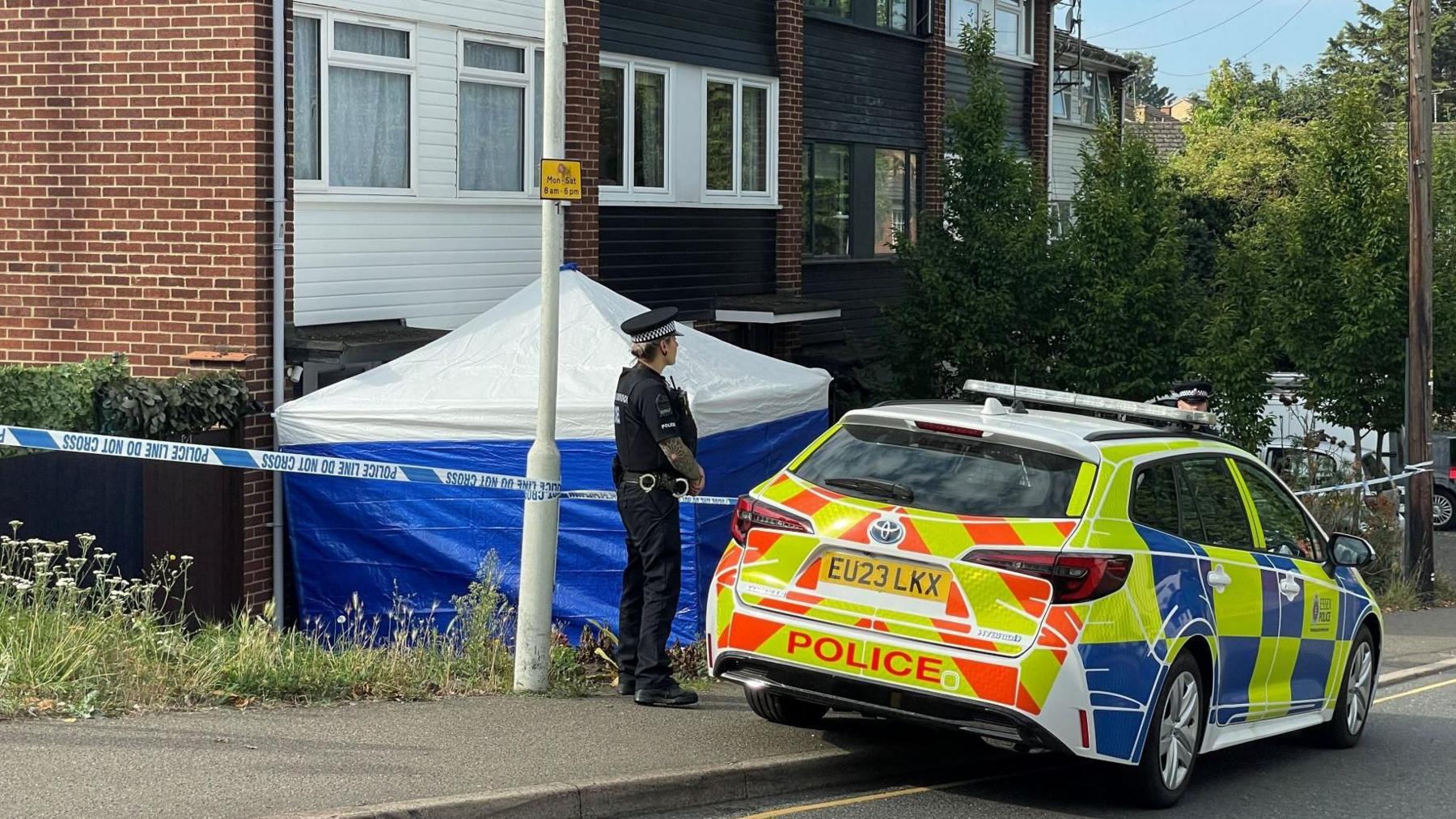 A police officer guarding the cordon at the McCullough property in Pump Hill. They have a police car in front of them and to their side is a blue sided police tent with a white top.