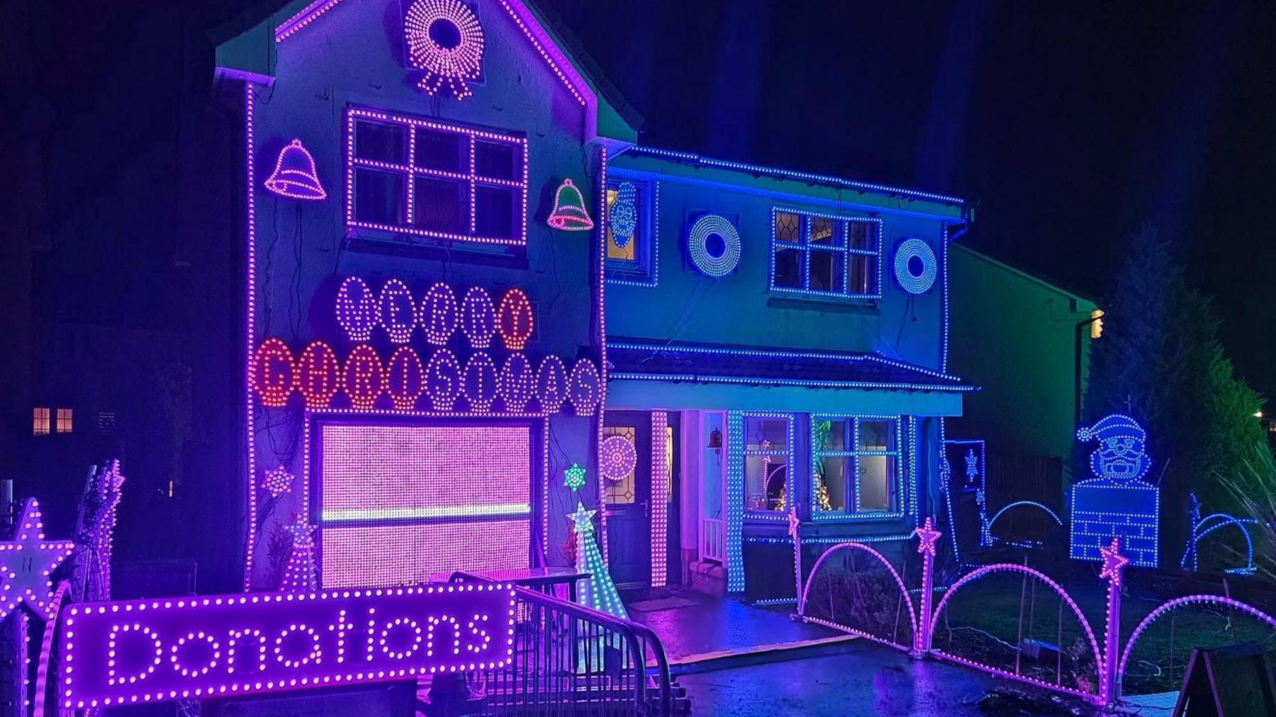 A two storey house brightly illuminated with LED Christmas lights in shades of blue and pink. There is a sign above the garage door that reads Merry Christmas. In the front corner of the garden is a pink sign which reads "Donations". 