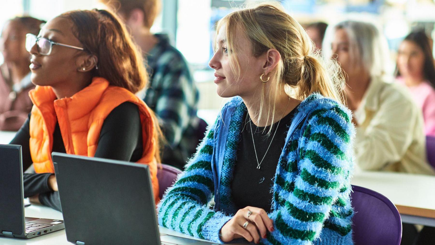 Two female students sit at a desk with laptops open. The student in the foreground on the right is wearing a bright blue cardigan over a black T-shirt, while the student on the left has glasses and a bright orange gilet. 