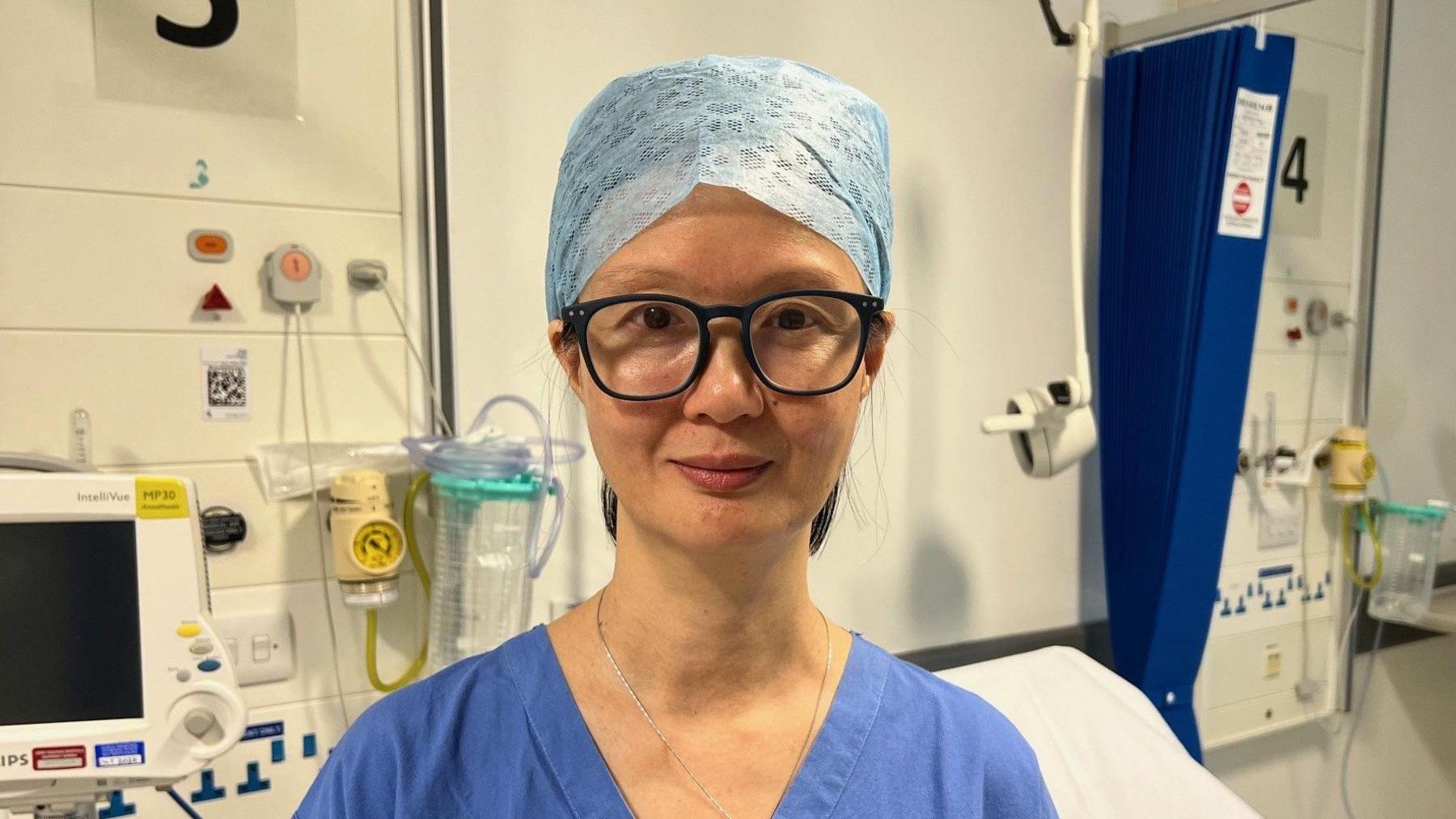 Prof Tze Min Wah stands in a hospital room. She is wearing blue scrubs and dark framed glasses.