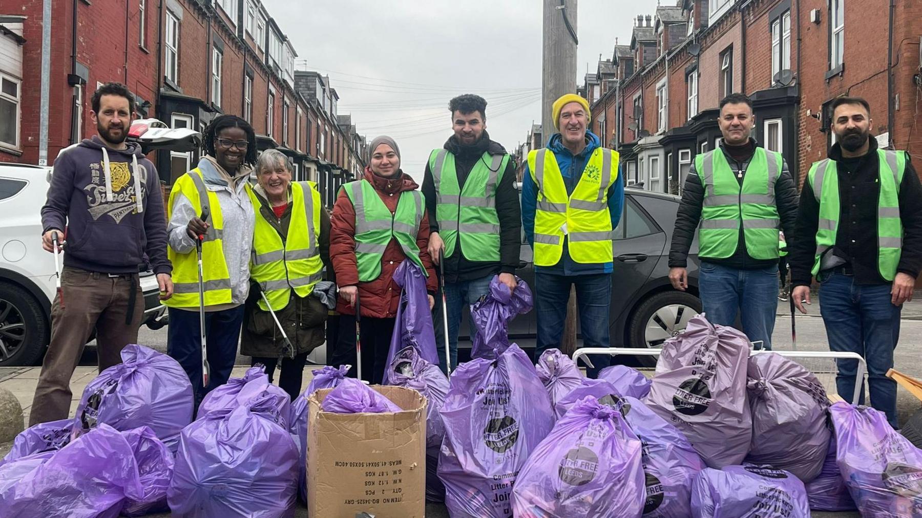  A group of men and women, most of them wearing luminous yellow or green bibs, stand on a street behind a collection of purple bags of rubbish. Terraced red-brick houses stand on either side of the street behind them.